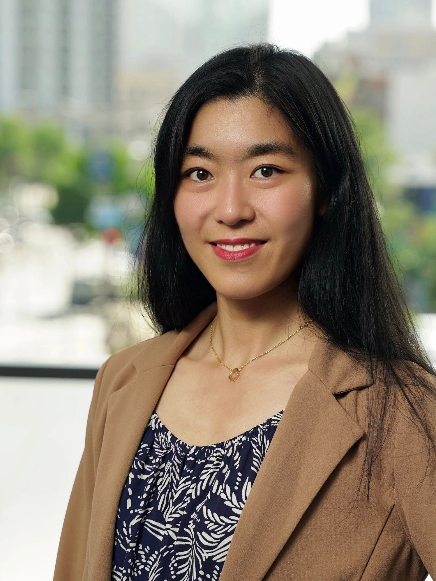A young woman with long black hair, wearing a tan blazer over a navy dress with white botanical print, and a gold necklace, standing indoors near a window with a cityscape background.