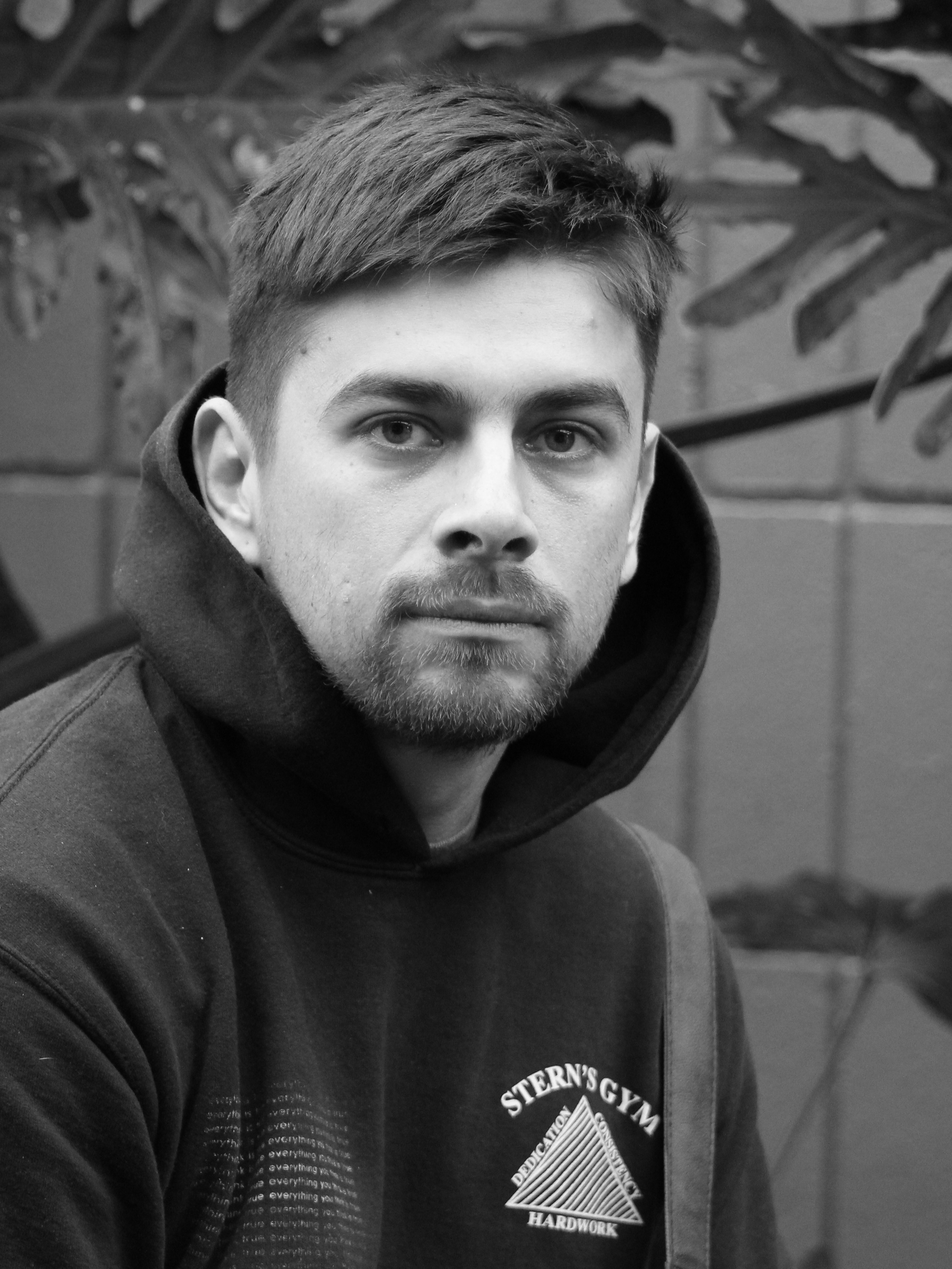 A young man with short hair and a beard wearing a hoodie with a gym logo on it, sitting indoors with a leafy background.