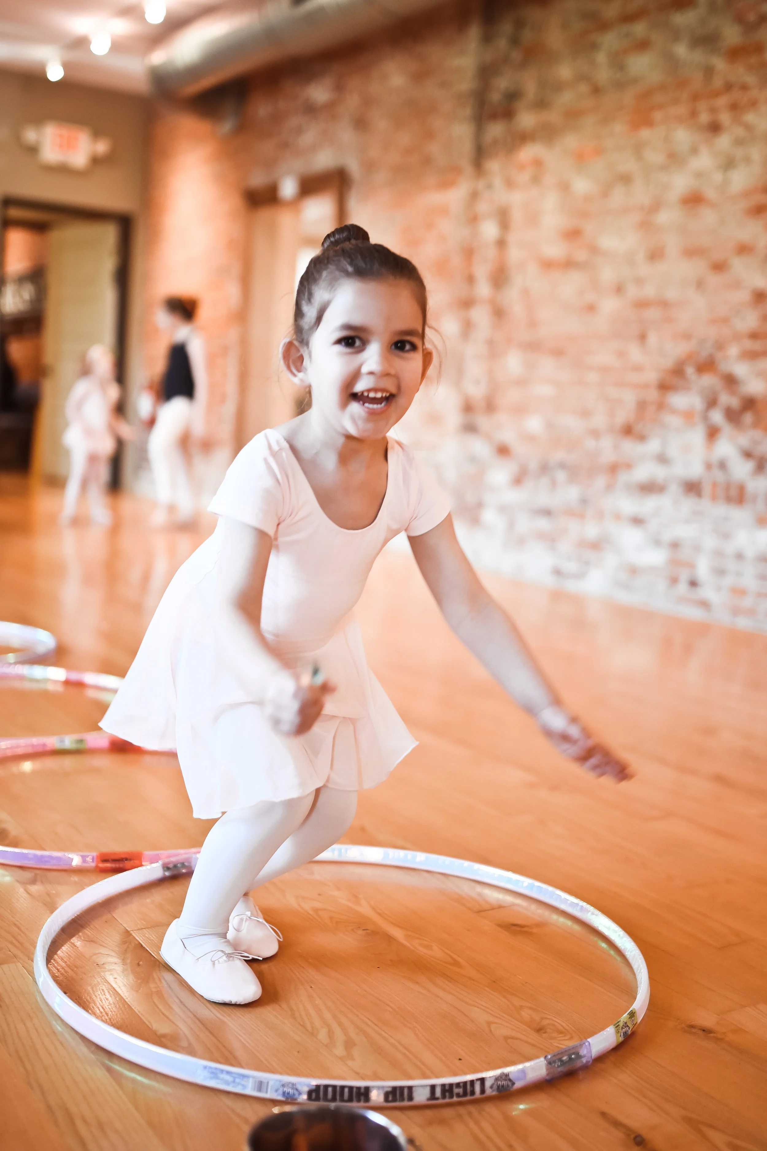 Young girl in a pink ballet outfit jumping inside a hula hoop in a dance studio with a brick wall background.