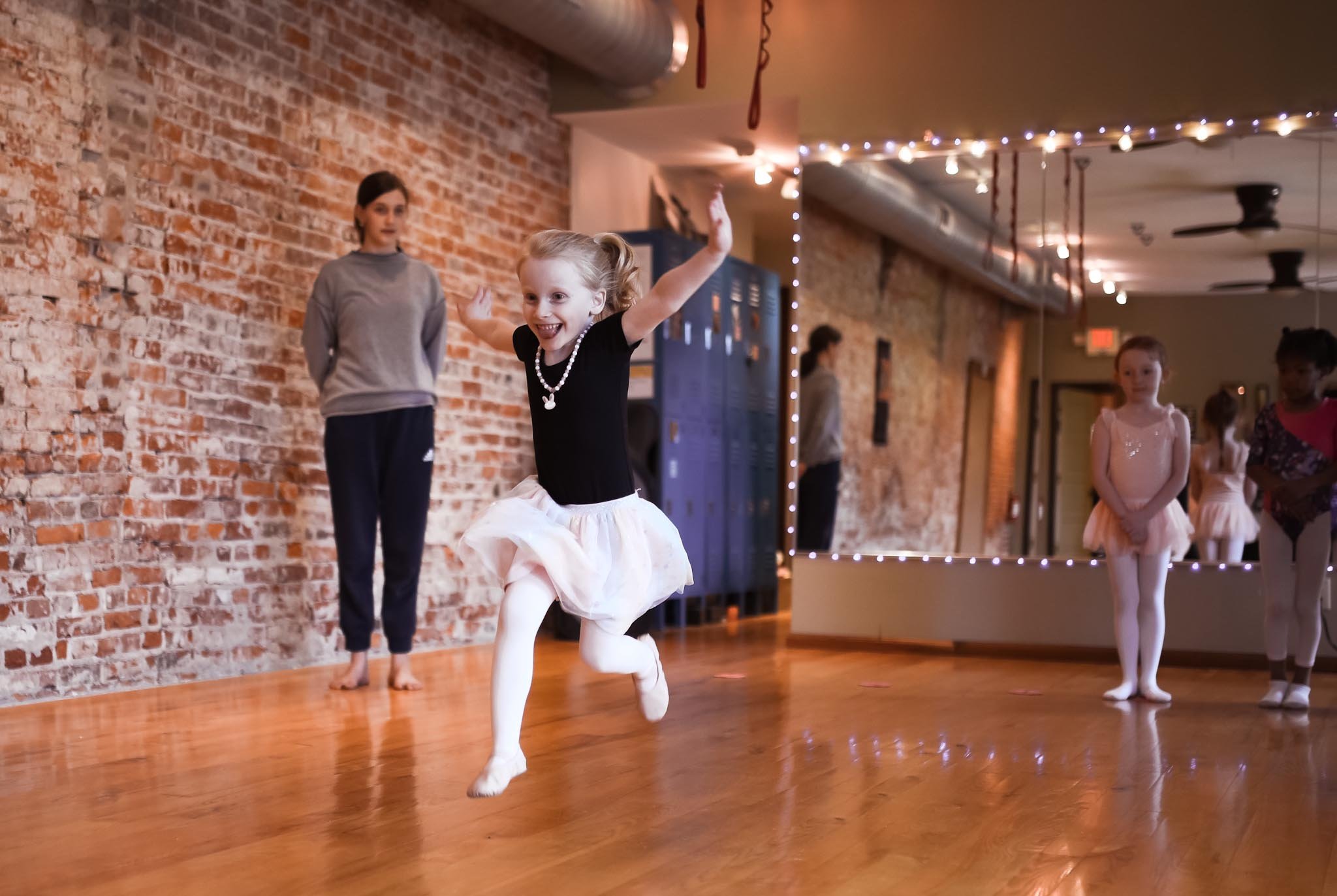 Two children, a boy and a girl, sitting on a wooden floor with their legs extended and arms raised, in front of an exposed brick wall.