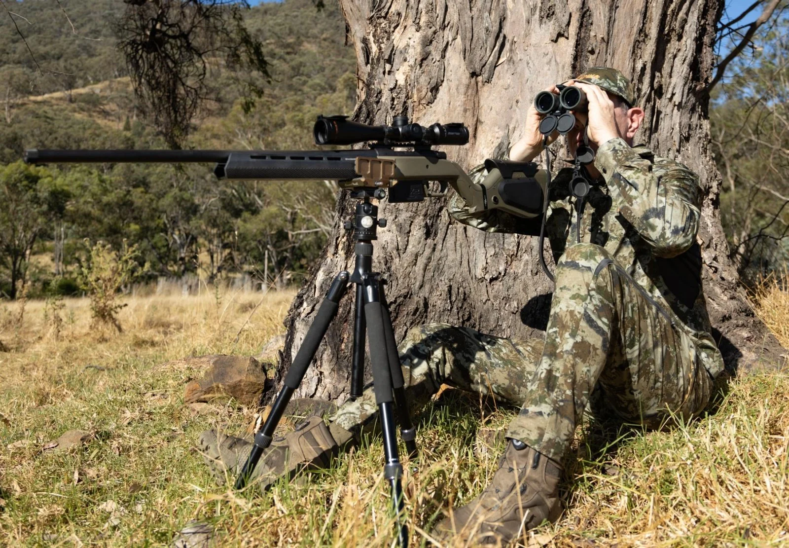 Camouflaged person sitting against a tree, holding binoculars, with a rifle on a tripod next to them in a woodland setting.