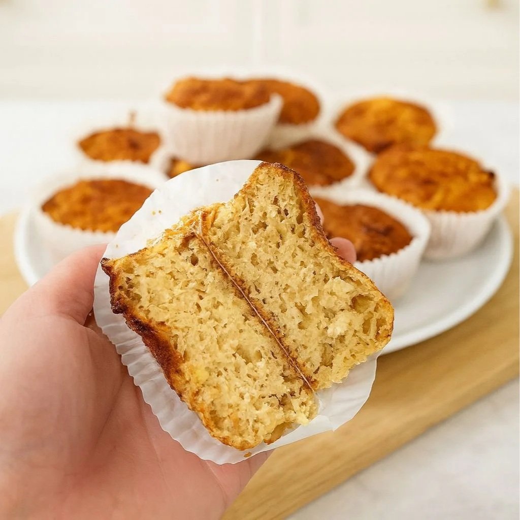 A hand holding a pineapple and cottage cheese muffin cut in half, showing the moist high-protein crumb with more muffins blurred in the background.