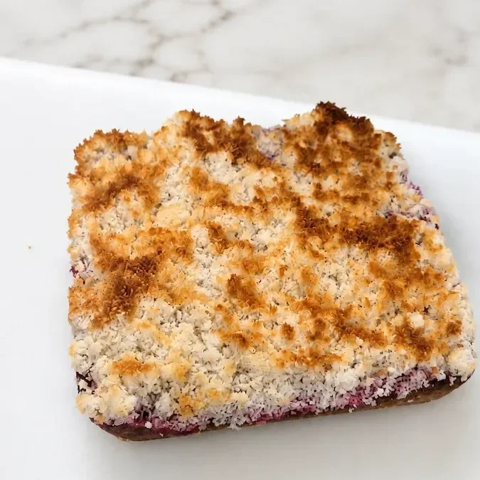 Whole uncut raspberry coconut slice with a golden toasted coconut topping on a white board over a marble bench.