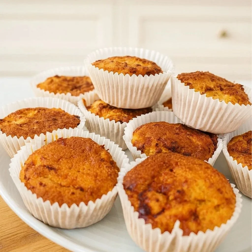 A plate of golden cottage cheese and pineapple muffins in white paper cases, sitting on a wooden chopping board with a bright kitchen background.