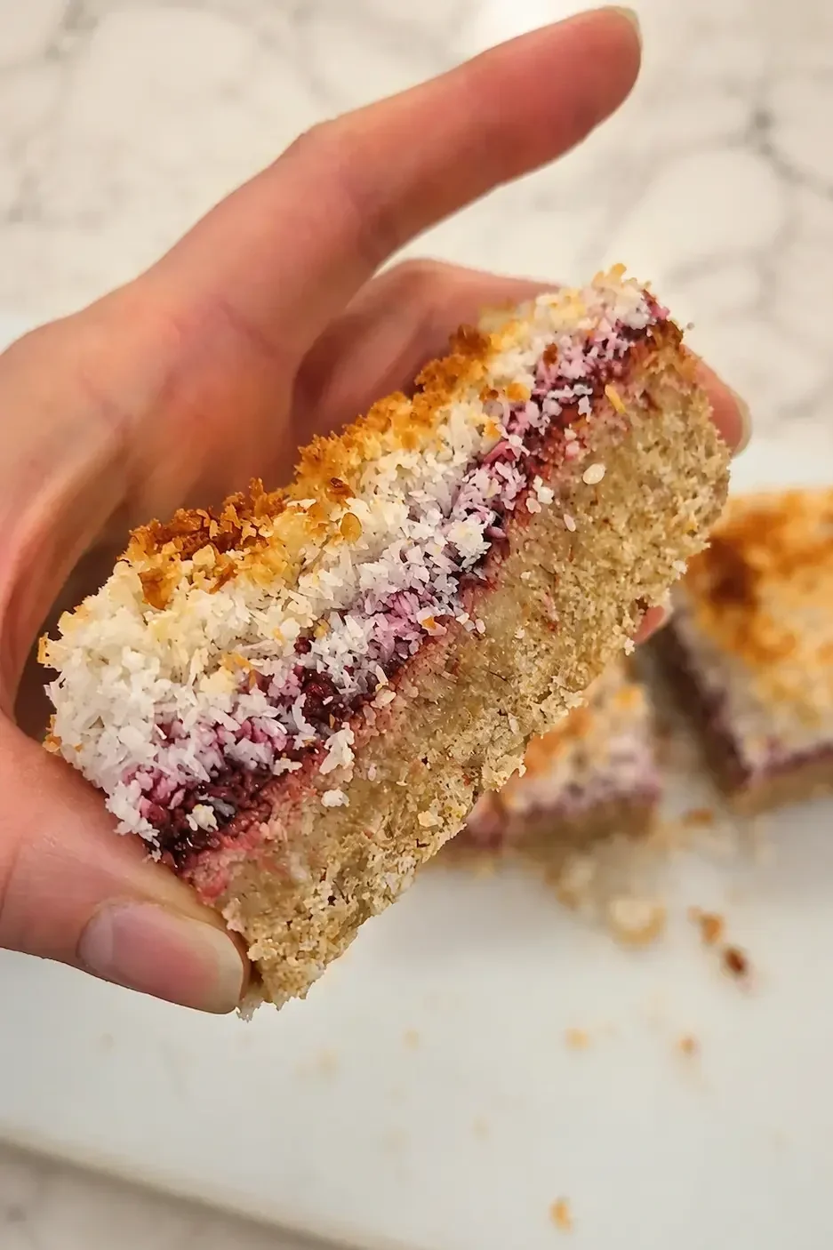 Hand holding a thick raspberry coconut slice with a flaxseed almond base, raspberry chia layer and toasted coconut topping.