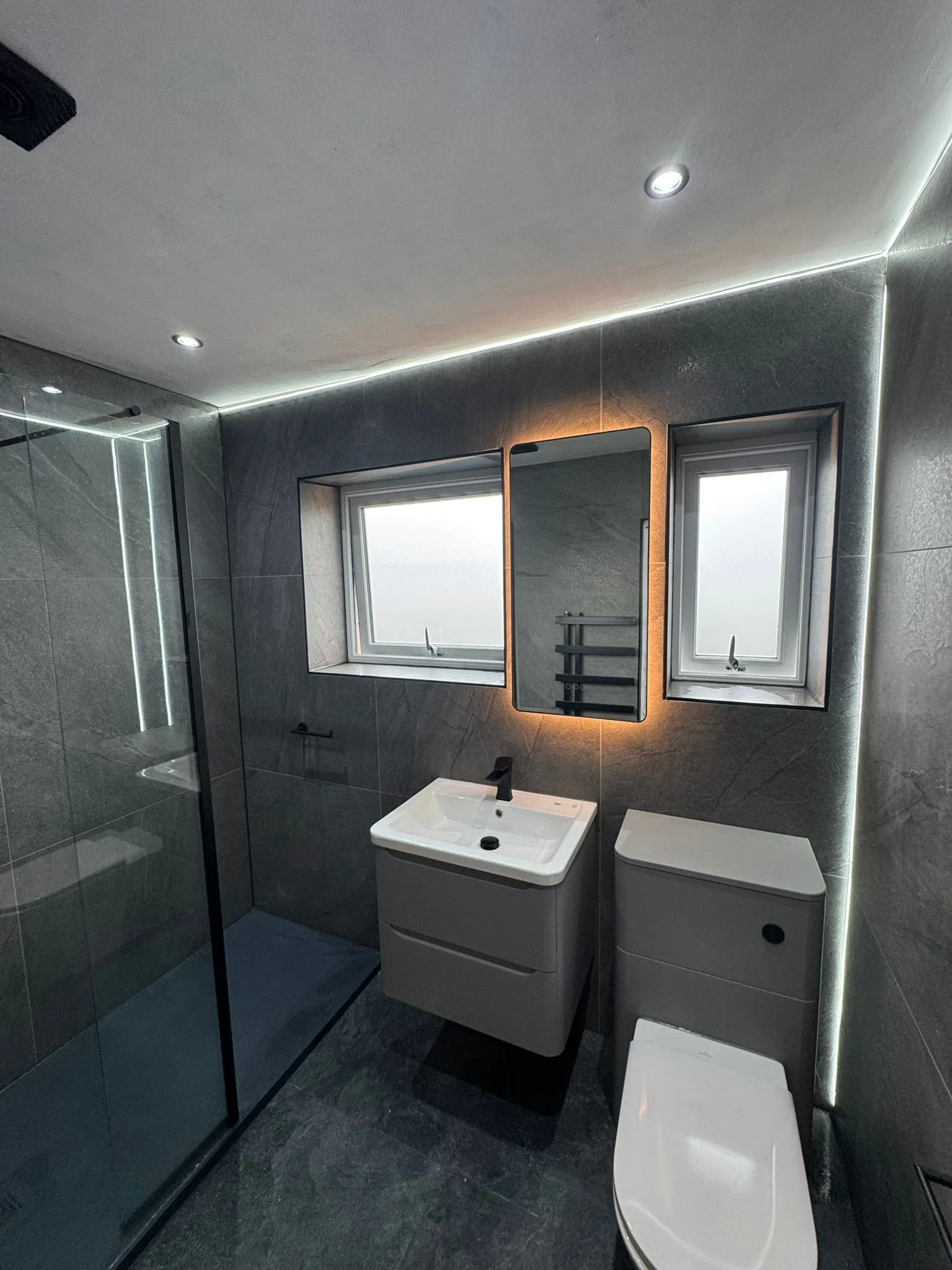 Modern bathroom in stanley with dark gray tiled walls and floor, two small frosted windows, a white sink with black faucet, rectangular mirror with backlight, a toilet, and a glass shower enclosure.