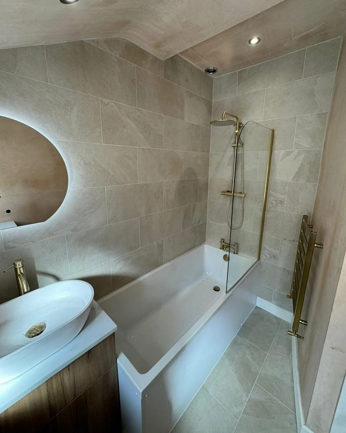 Modern bathroom in whitley bay with beige tiled walls and floor, a bathtub with a glass shower panel, a vessel sink with a brass faucet, a mirror, and a heated towel rack.