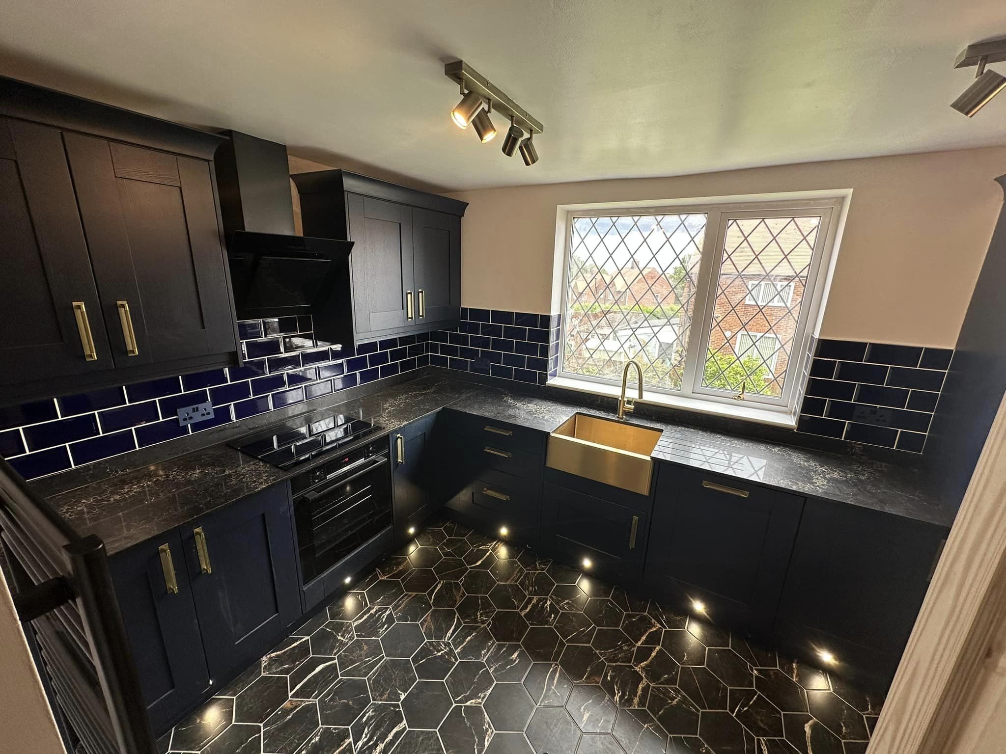 Modern kitchen in sunderland with black cabinets, black marble countertops, a gold-colored belfast sink, navy blue subway tile backsplash, and black hexagon tile flooring with white veining. 