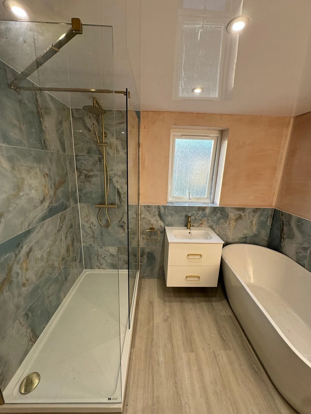 Modern bathroom in durham with a large blue  marble-tiled shower, a white vanity with gold handles, a freestanding bathtub, a small window, and ceiling lights.