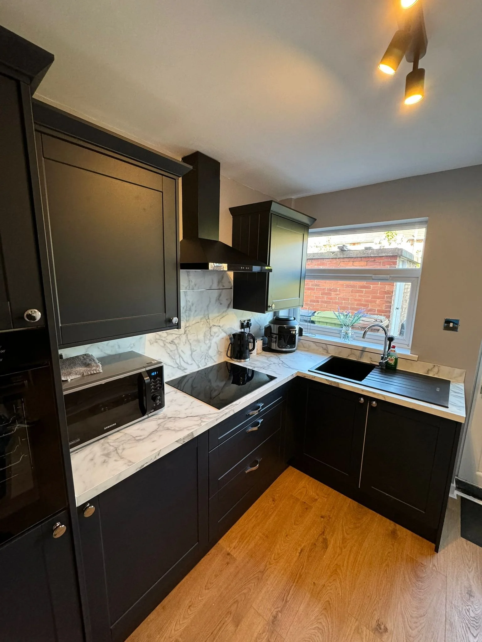 Kitchen with black cabinets, marble countertops, a window, and wooden flooring.