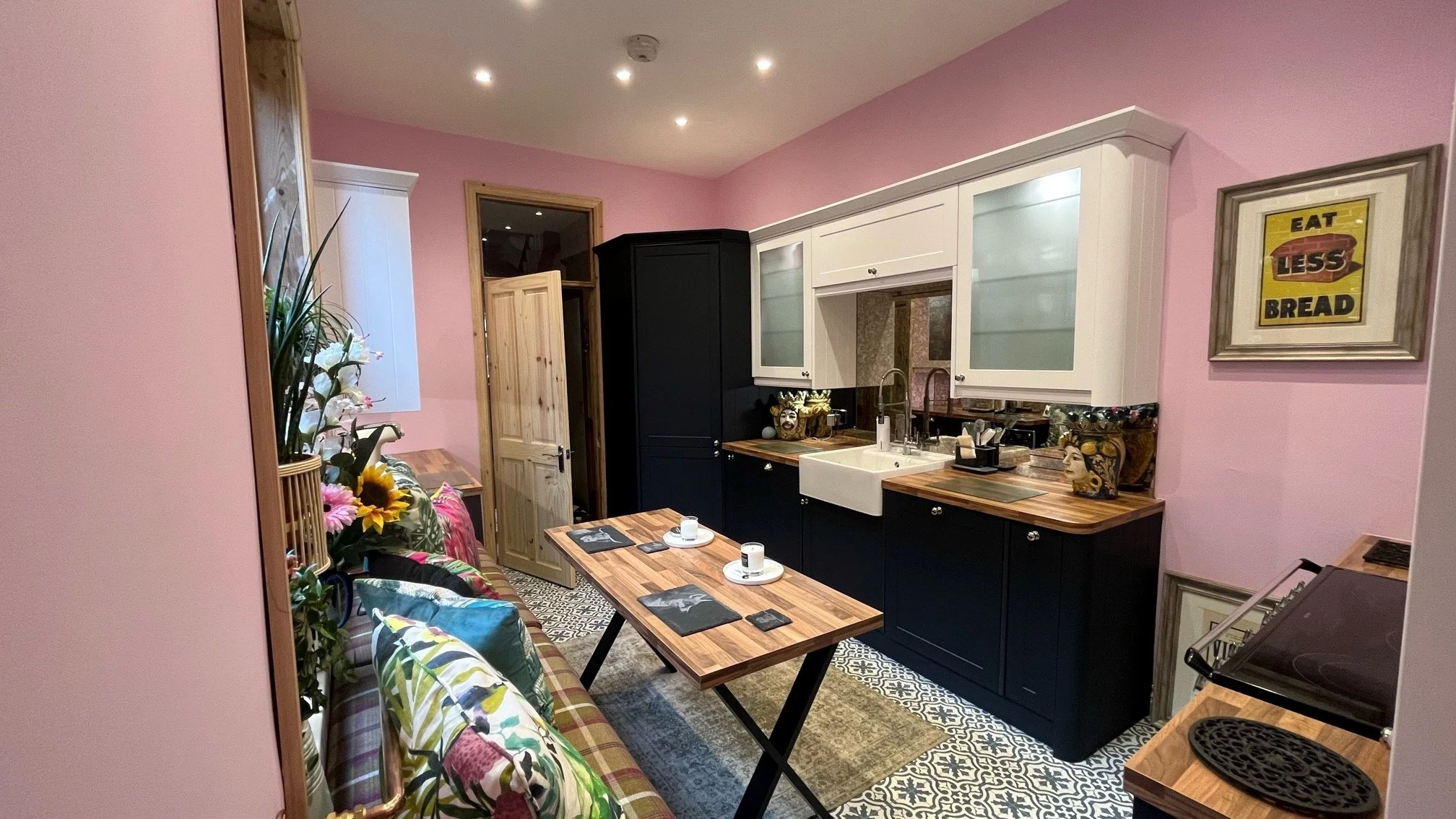 A cozy kitchen and dining area in sunderland  with pink walls, black and white cabinetry, a belfast sink, and a wooden table set for coffee. Decorative vases, framed artwork, and patterned flooring complete the space.