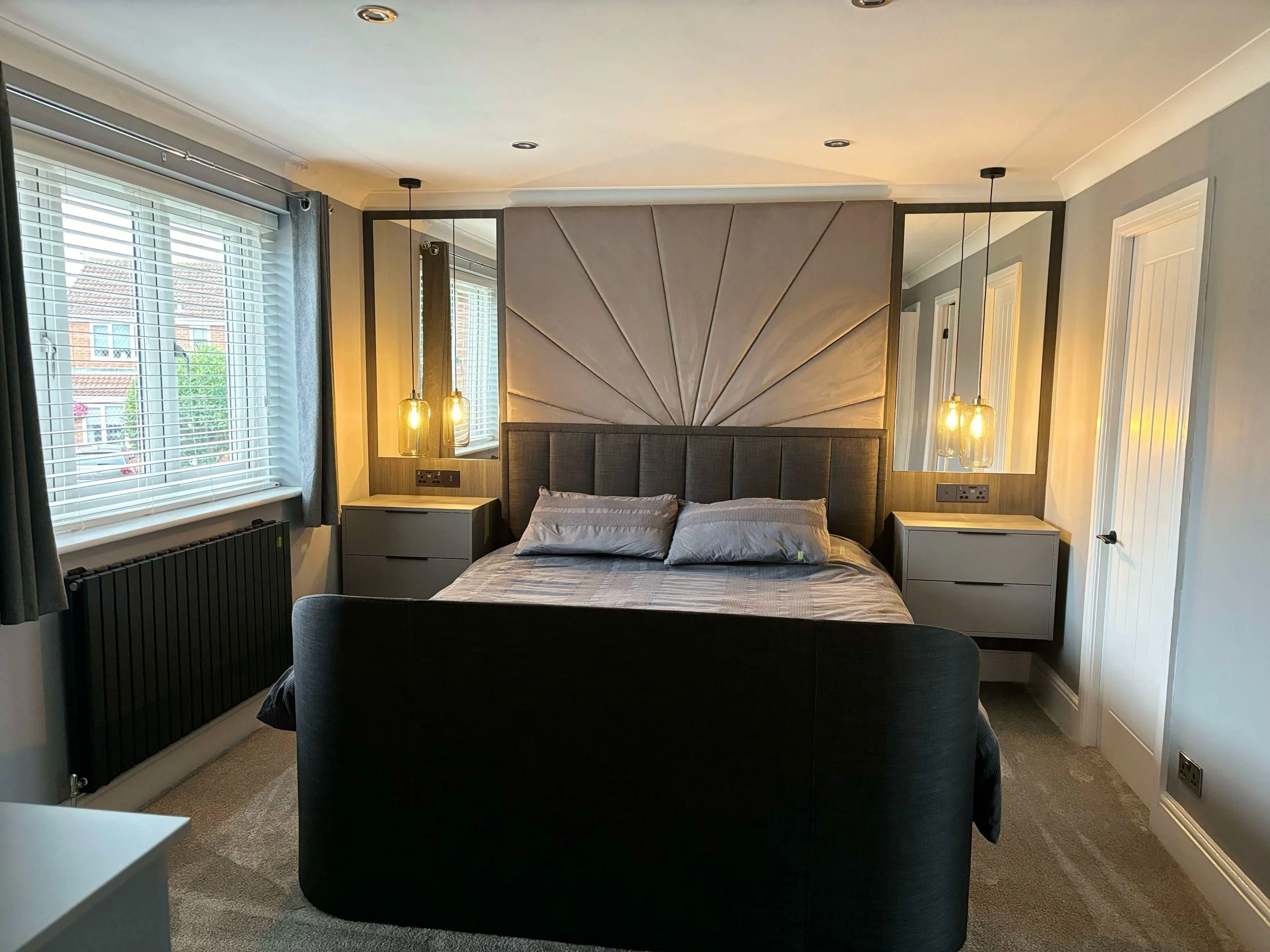 Modern bedroom with a bed, two nightstands, pendant lighting, windows with blinds, and a geometric headboard design.