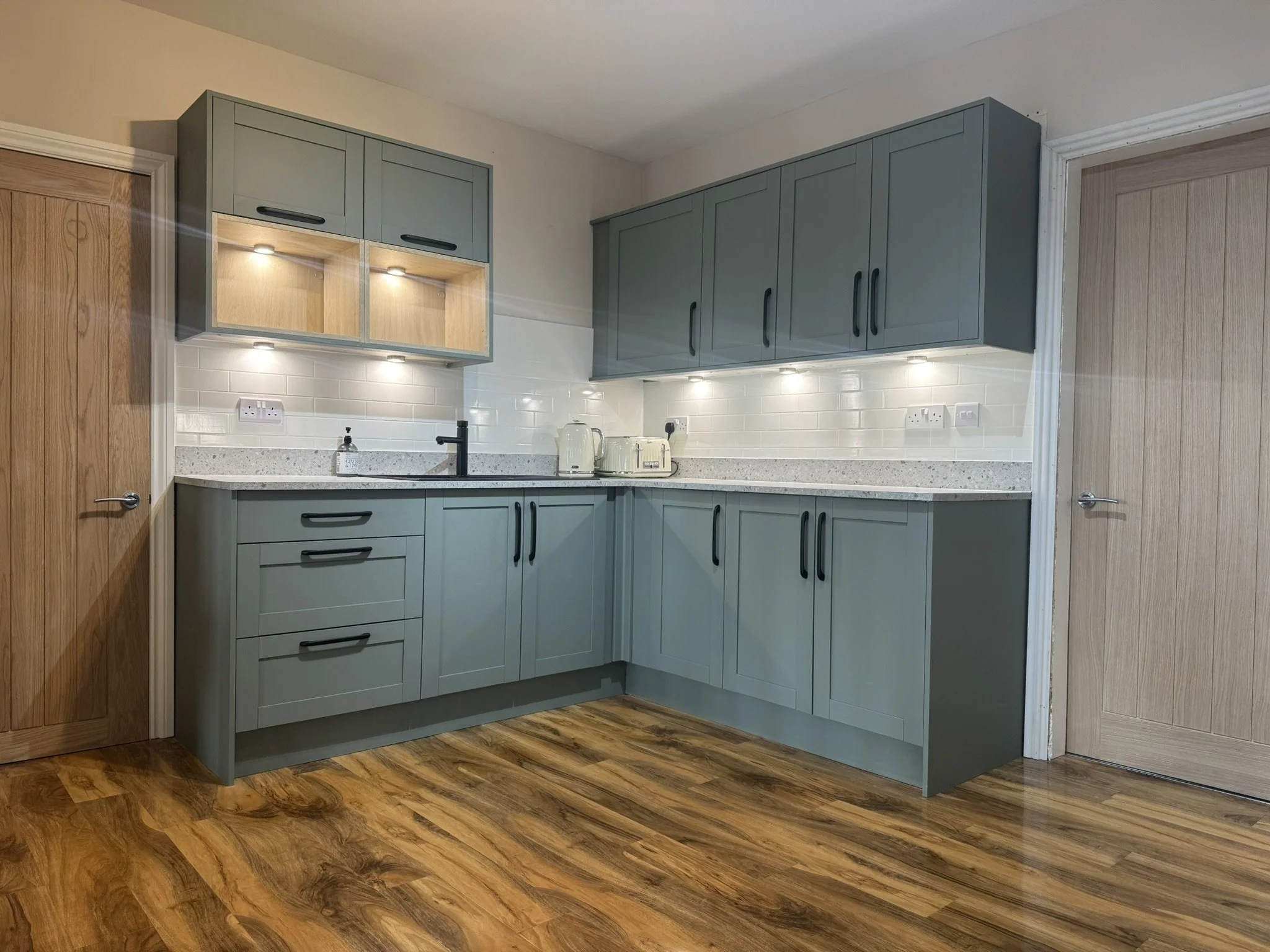 Modern kitchen with grey cabinets, white backsplash, black faucet, and wooden flooring.