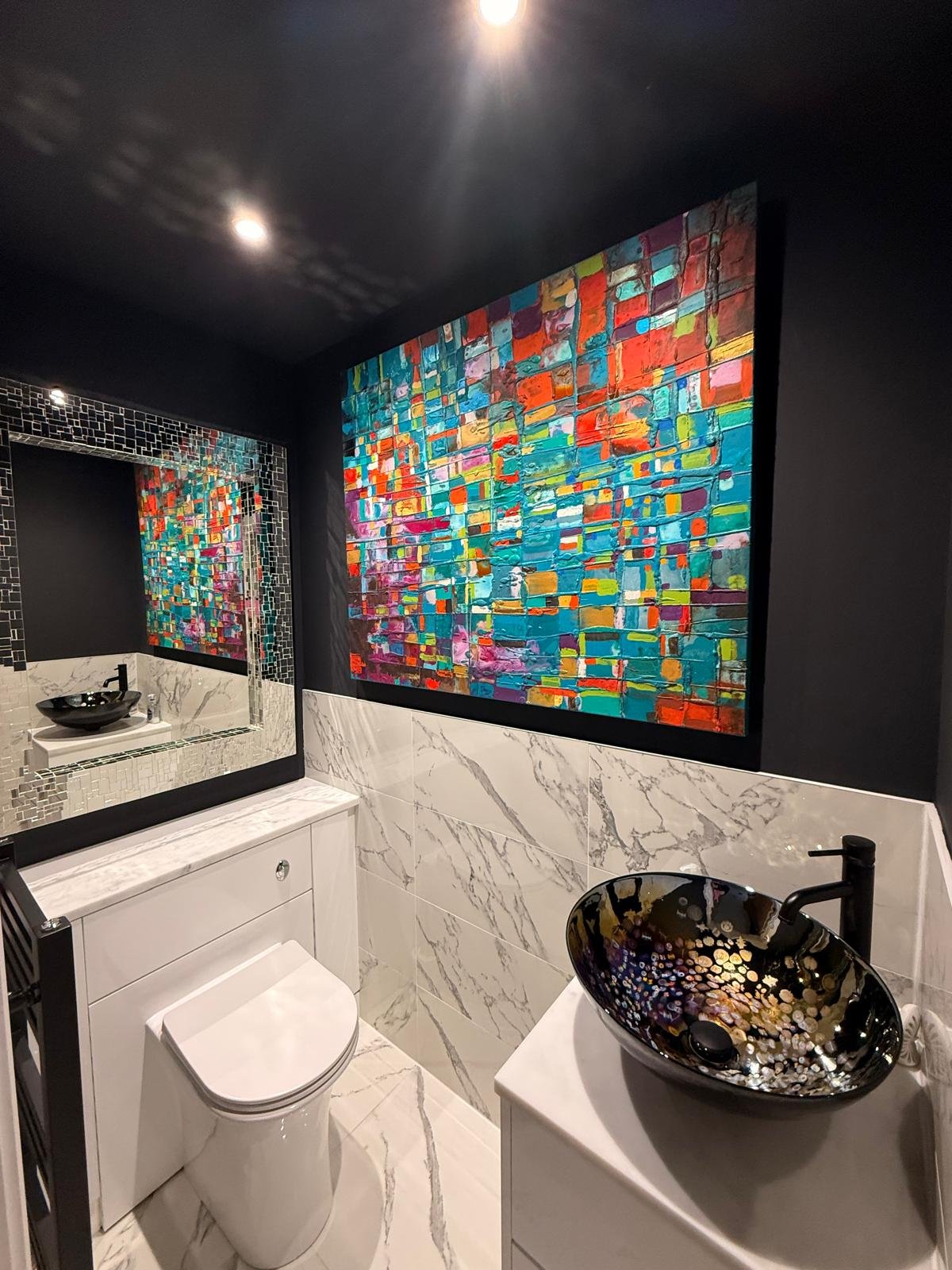 A small modern bathroom in morpeth with black walls and marble tile accents. Features a colorful abstract painting on the wall, a large mirror with a mosaic border, a black vessel sink on a white cabinet, and a toilet.