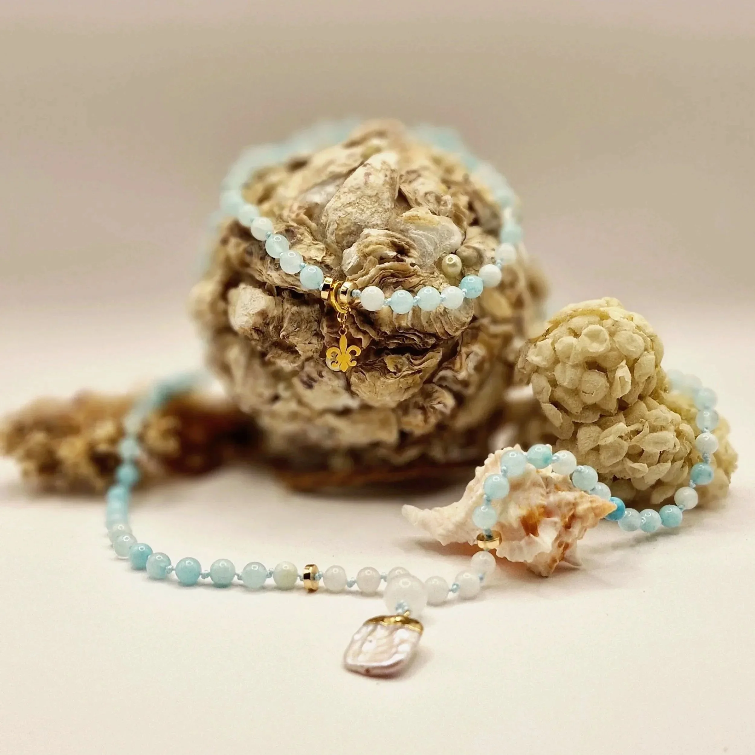 Kette aus hellblauen Natursteinperlen mit Süßwasserperle als Anhänger und goldfarbenen Akzenten dekoriert mit Muscheln auf hellem Untergrund