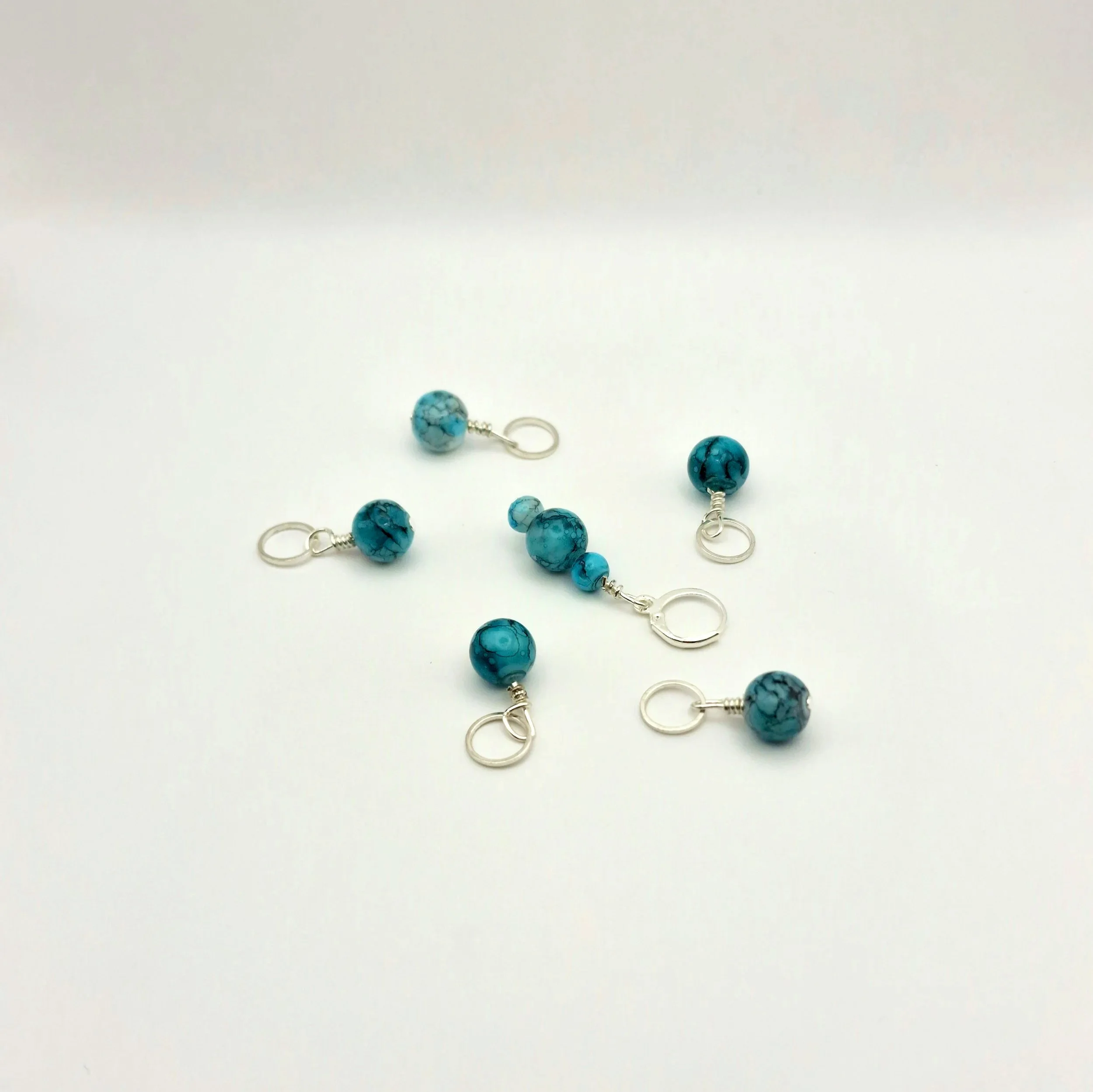 Set aus fünf Maschenmarkierern und einem Progresskeeper aus Teal und türkisfarbenen handbemalten Keramikperlen kombiniert mit versilberten Ringen auf weißem Untergrund