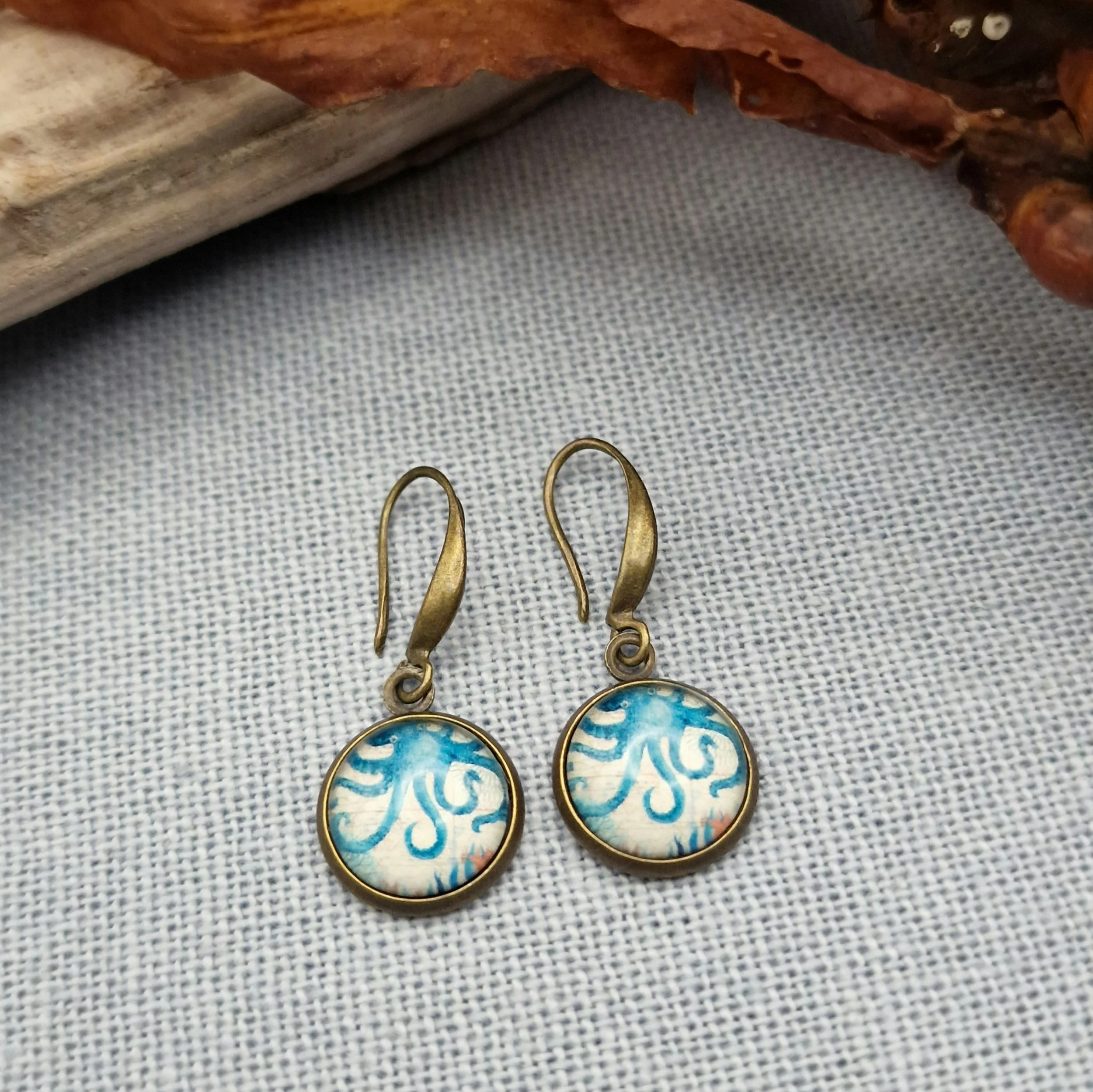 hängende Ohrringe aus Bronzefarbenem Metall mit Octopus Cabochon aus Glas in maritimen Vintage Stil liegend auf hellblauem Untergrund mit Dekoration aus getrocknete Seetang und Treibholz