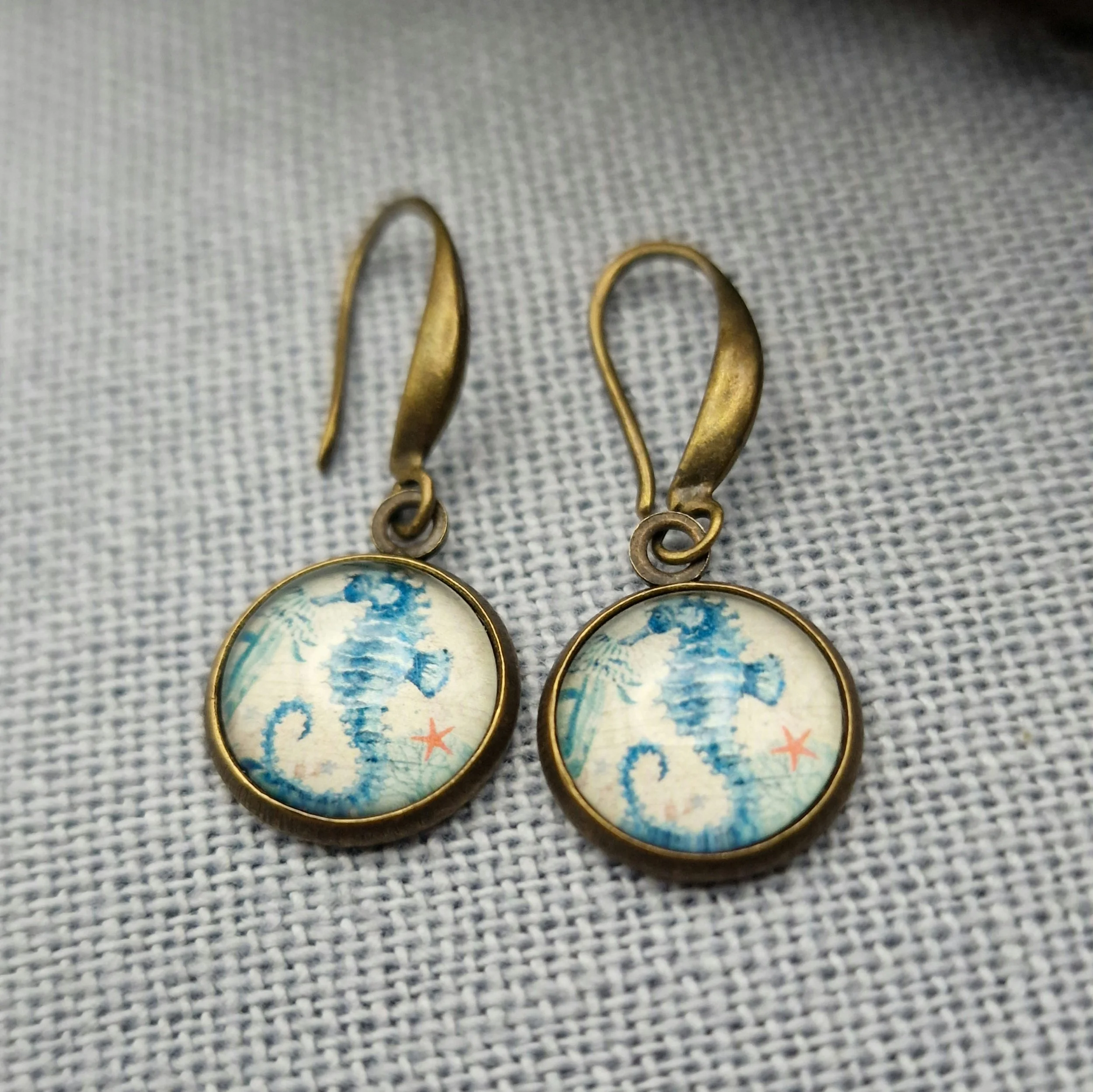 ein paar hängende Ohrringe in Bronze mit Cabochon mit Bild von Seepferdchen liegen auf auf hellblauem Untergrund