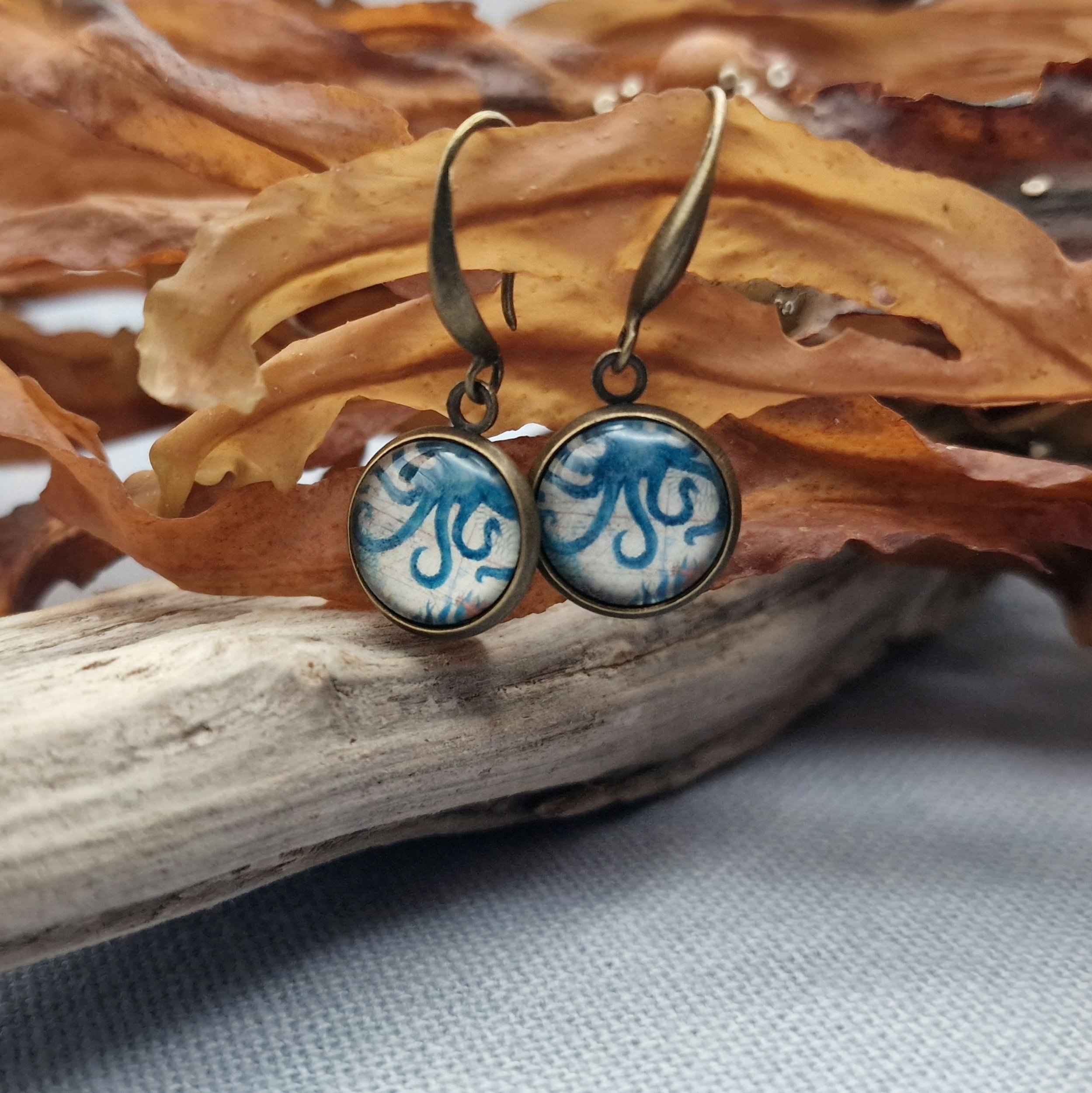 hängende Ohrringe aus Bronzefarbenem Metall mit Octopus Cabochon aus Glas in maritimen Vintage Stil liegend auf hellblauem Untergrund mit Dekoration aus getrocknete Seetang und Treibholz