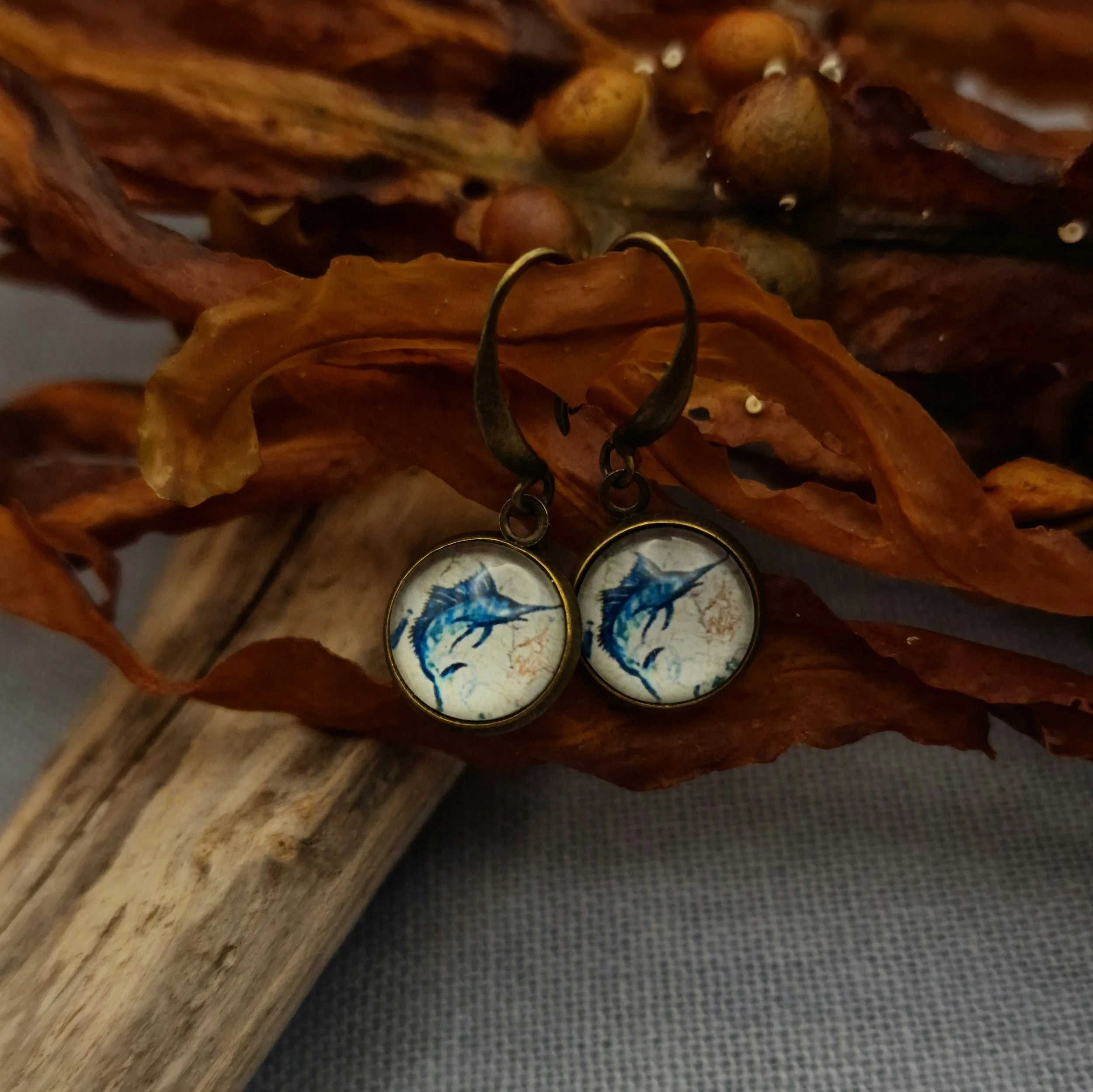 ein paar hängende Ohrringe in Bronze mit Cabochon mit Bild von Schwertfisch hängen an getrocknetem Seetang und Treibholz auf hellblauem Untergrund