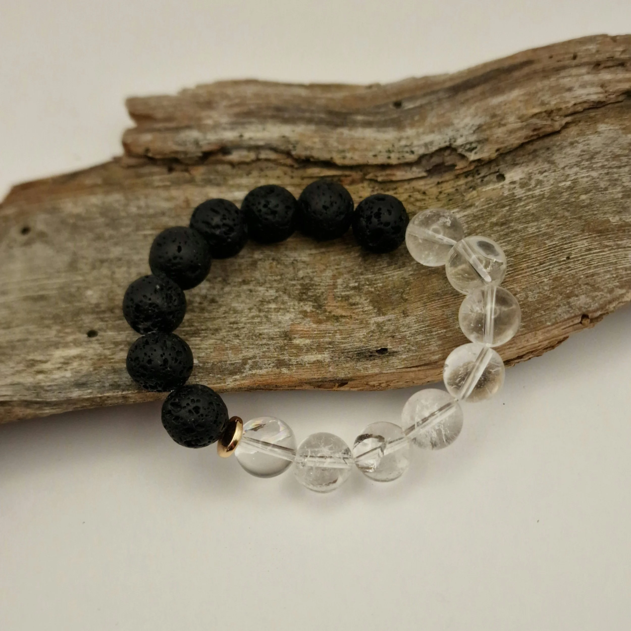 Armband aus schwarzen Lavaperlen und klarem Bergkristall mit einer vergoldeten Hämatit-Disc auf einem Stück Treibholz mit hellem Untergrund