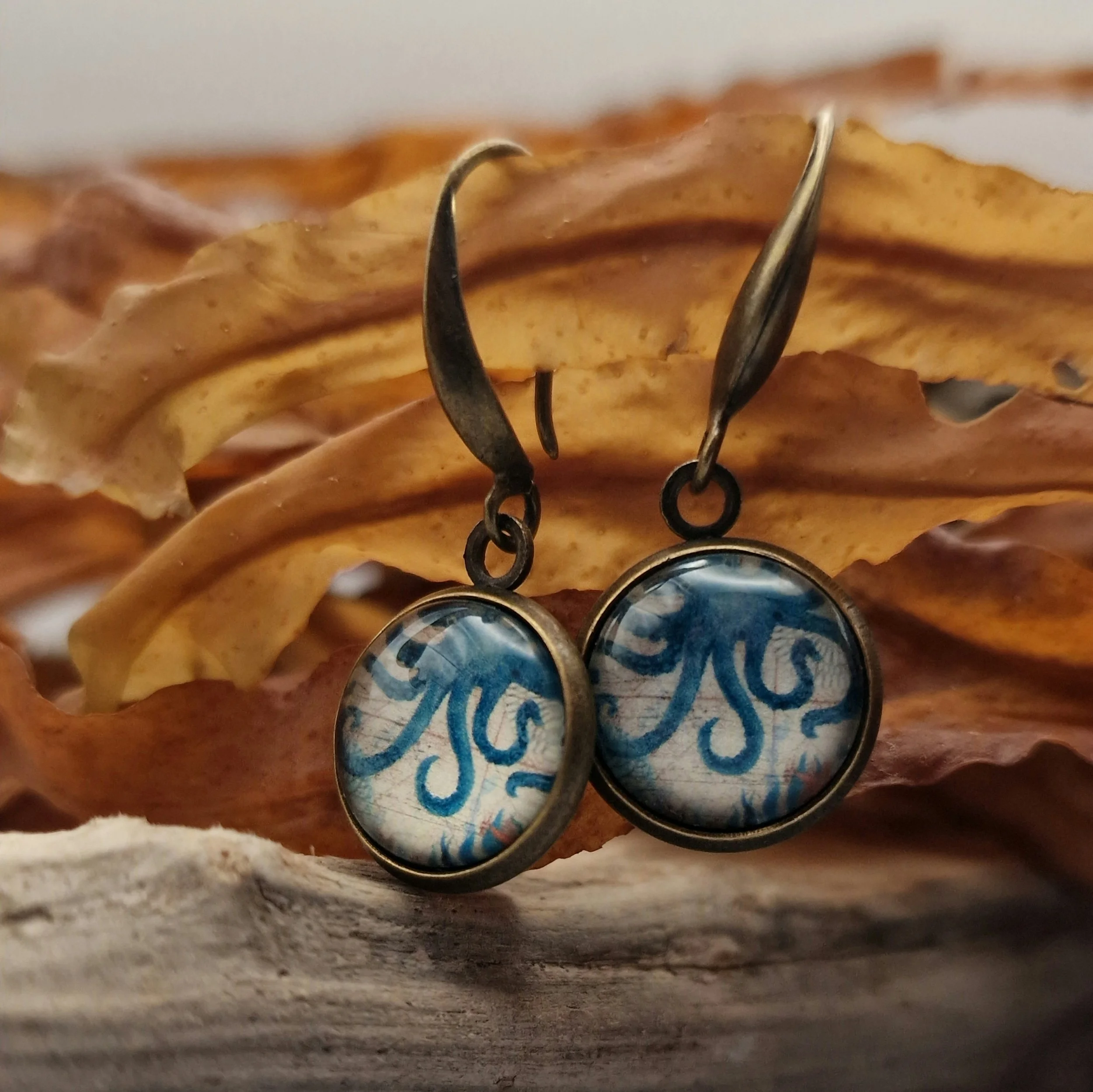hängende Ohrringe aus Bronzefarbenem Metall mit Octopus Cabochon aus Glas in maritimen Vintage Stil liegend auf hellblauem Untergrund mit Dekoration aus getrocknete Seetang und Treibholz