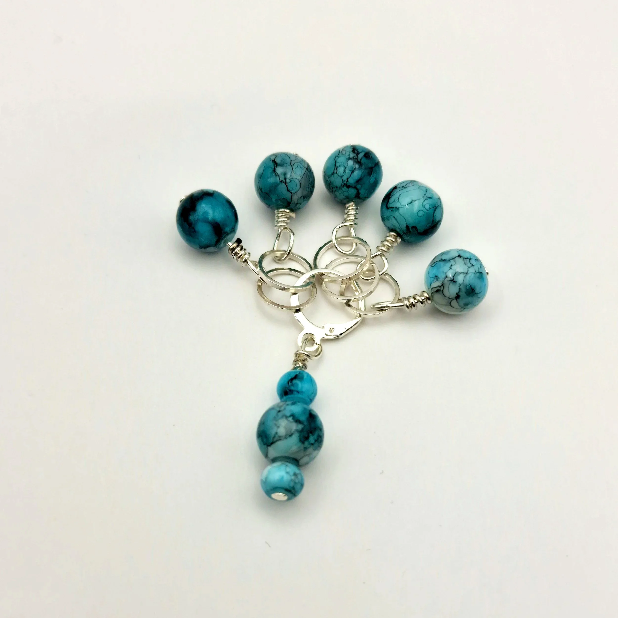 Set aus fünf Maschenmarkierern und einem Progresskeeper aus Teal und türkisfarbenen handbemalten Keramikperlen kombiniert mit versilberten Ringen auf weißem Untergrund
