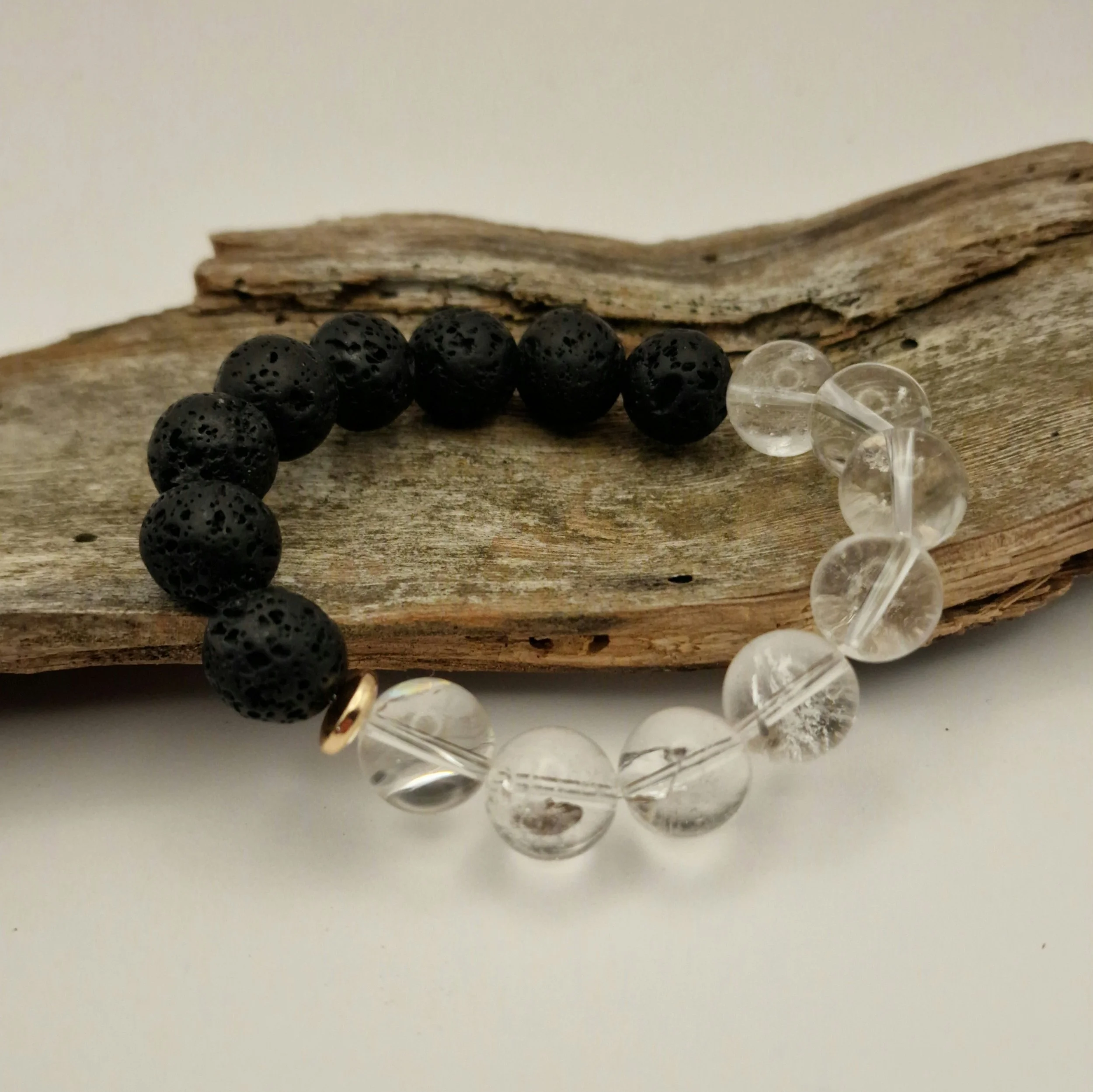Armband aus schwarzen Lavaperlen und klarem Bergkristall mit einer vergoldeten Hämatit-Disc auf einem Stück Treibholz mit hellem Untergrund