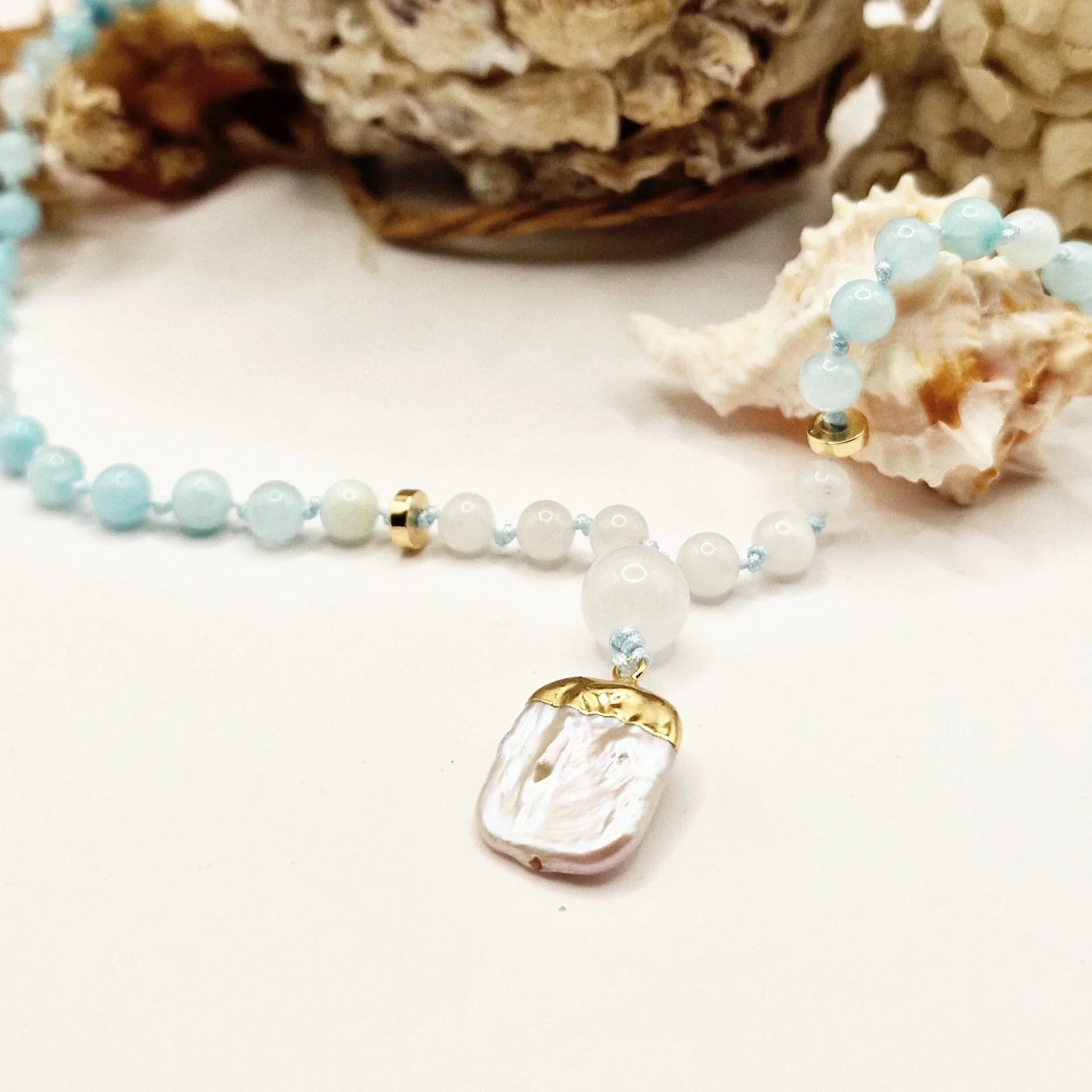 Kette aus hellblauen Natursteinperlen mit Süßwasserperle als Anhänger und goldfarbenen Akzenten dekoriert mit Muscheln auf hellem Untergrund