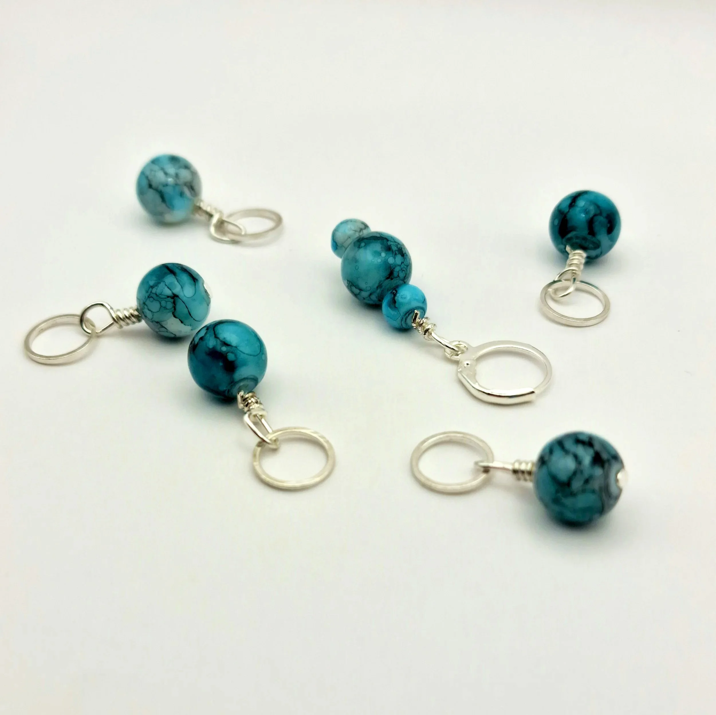 Set aus fünf Maschenmarkierern und einem Progresskeeper aus Teal und türkisfarbenen handbemalten Keramikperlen kombiniert mit versilberten Ringen auf weißem Untergrund