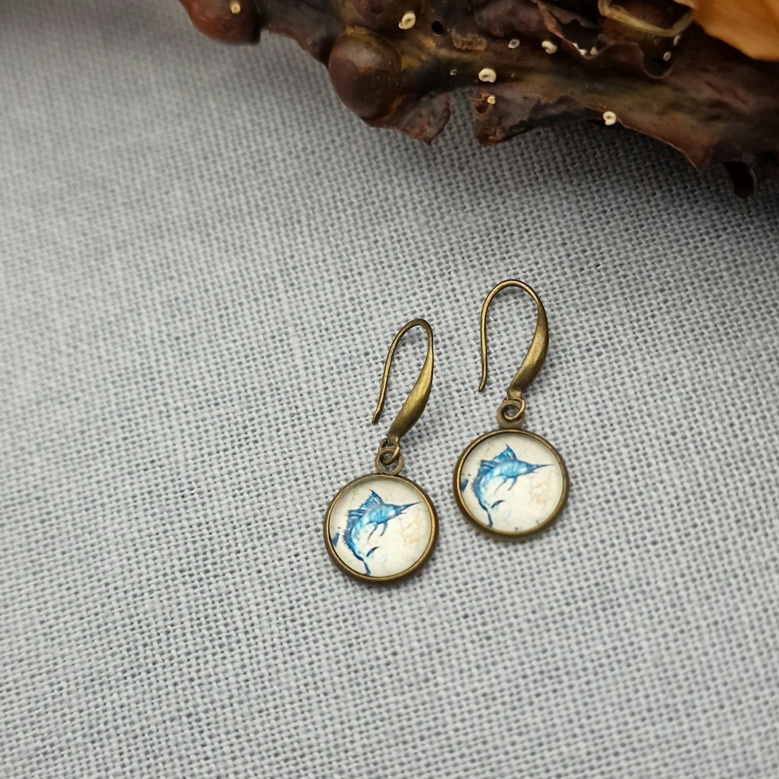 ein paar hängende Ohrringe in Bronze mit Cabochon mit Bild von Schwertfisch hängen an getrocknetem Seetang und Treibholz auf hellblauem Untergrund