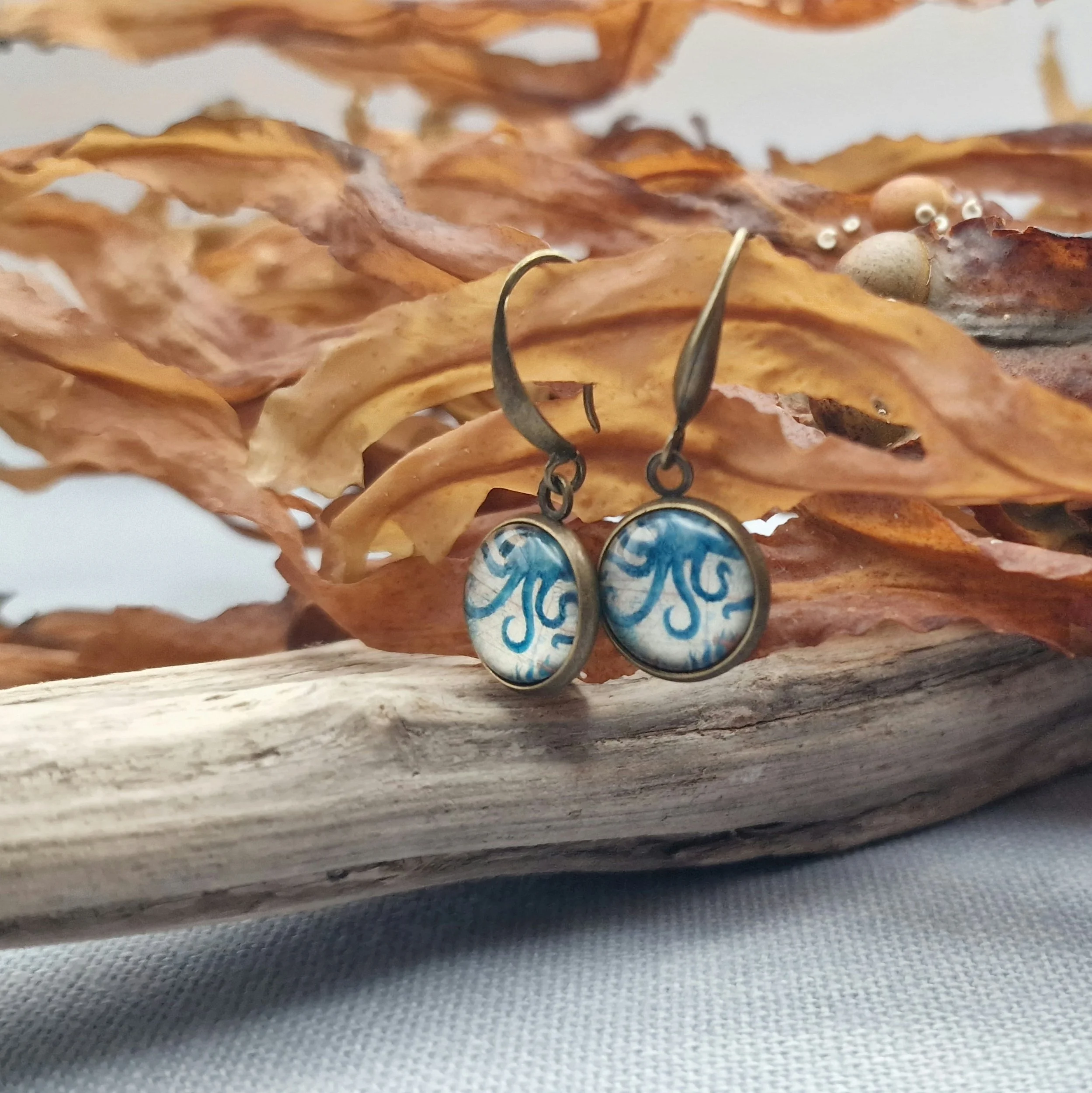 hängende Ohrringe aus Bronzefarbenem Metall mit Octopus Cabochon aus Glas in maritimen Vintage Stil liegend auf hellblauem Untergrund mit Dekoration aus getrocknete Seetang und Treibholz