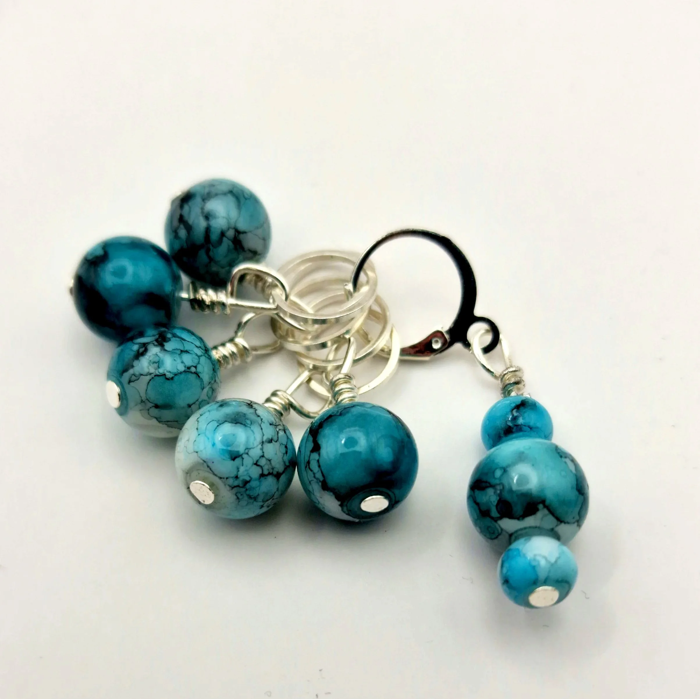 Set aus fünf Maschenmarkierern und einem Progresskeeper aus Teal und türkisfarbenen handbemalten Keramikperlen kombiniert mit versilberten Ringen auf weißem Untergrund