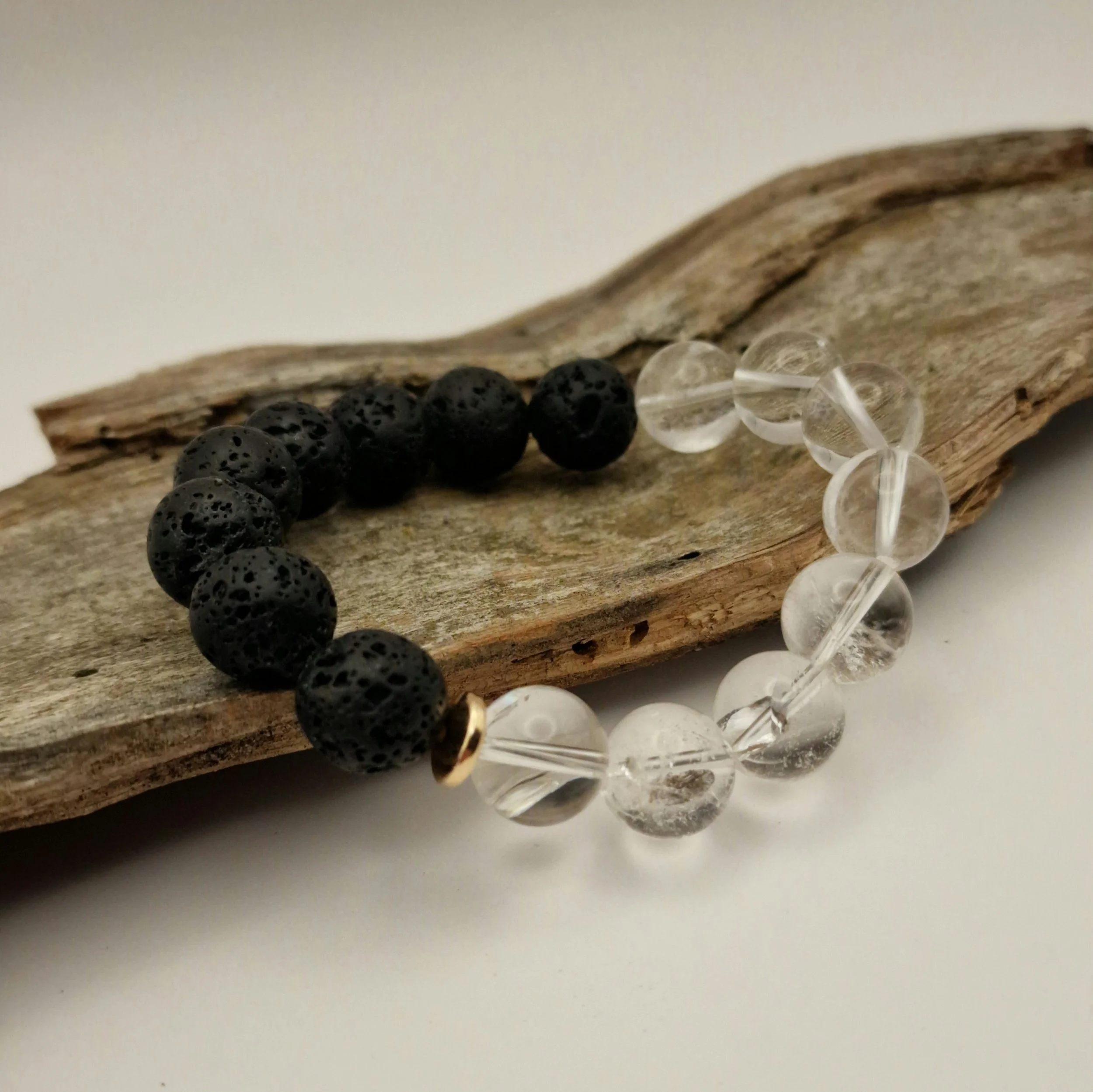 Armband aus schwarzen Lavaperlen und klarem Bergkristall mit einer vergoldeten Hämatit-Disc auf einem Stück Treibholz mit hellem Untergrund