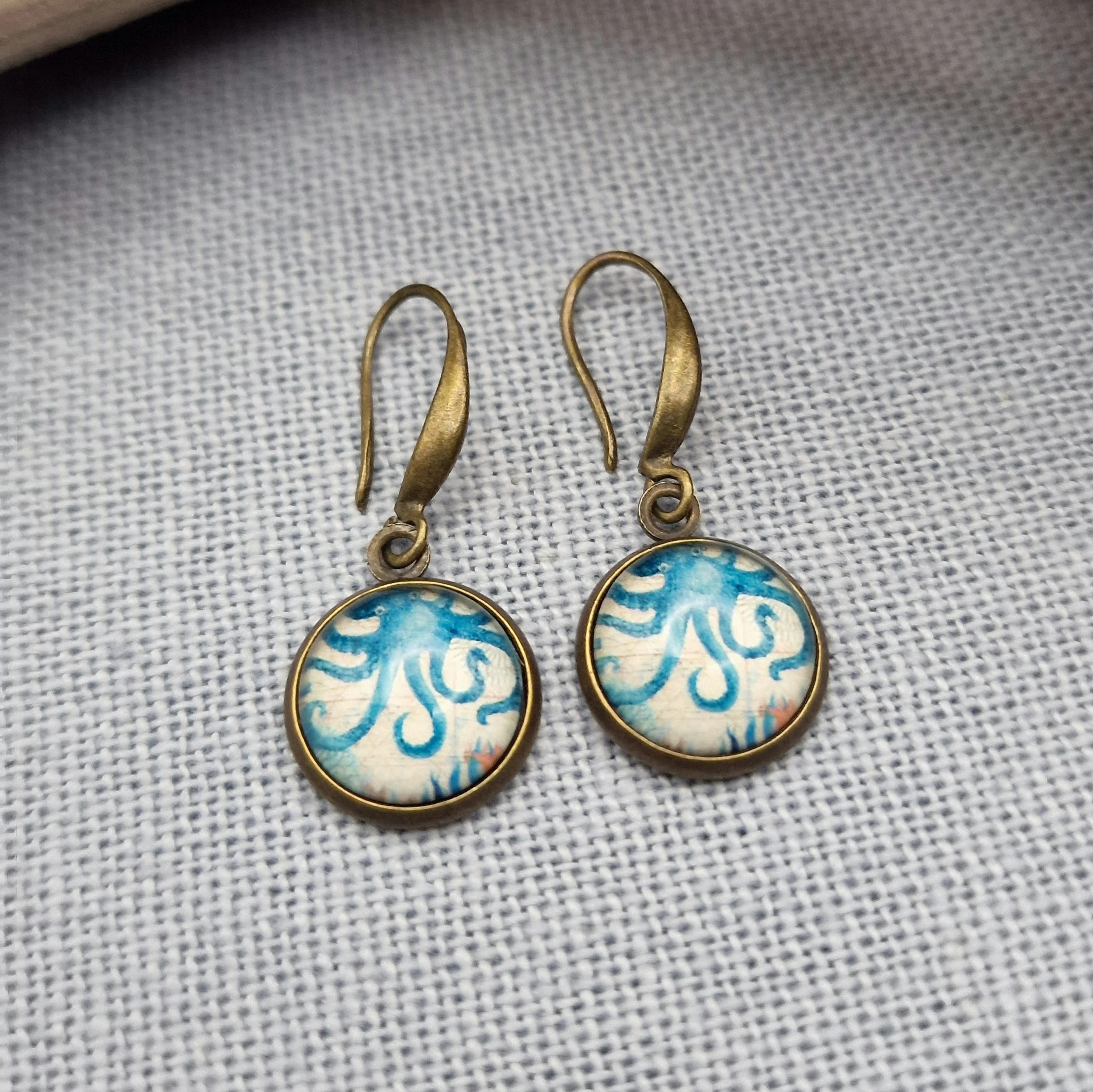 hängende Ohrringe aus Bronzefarbenem Metall mit Octopus Cabochon aus Glas in maritimen Vintage Stil liegend auf hellblauem Untergrund mit Dekoration aus getrocknete Seetang und Treibholz