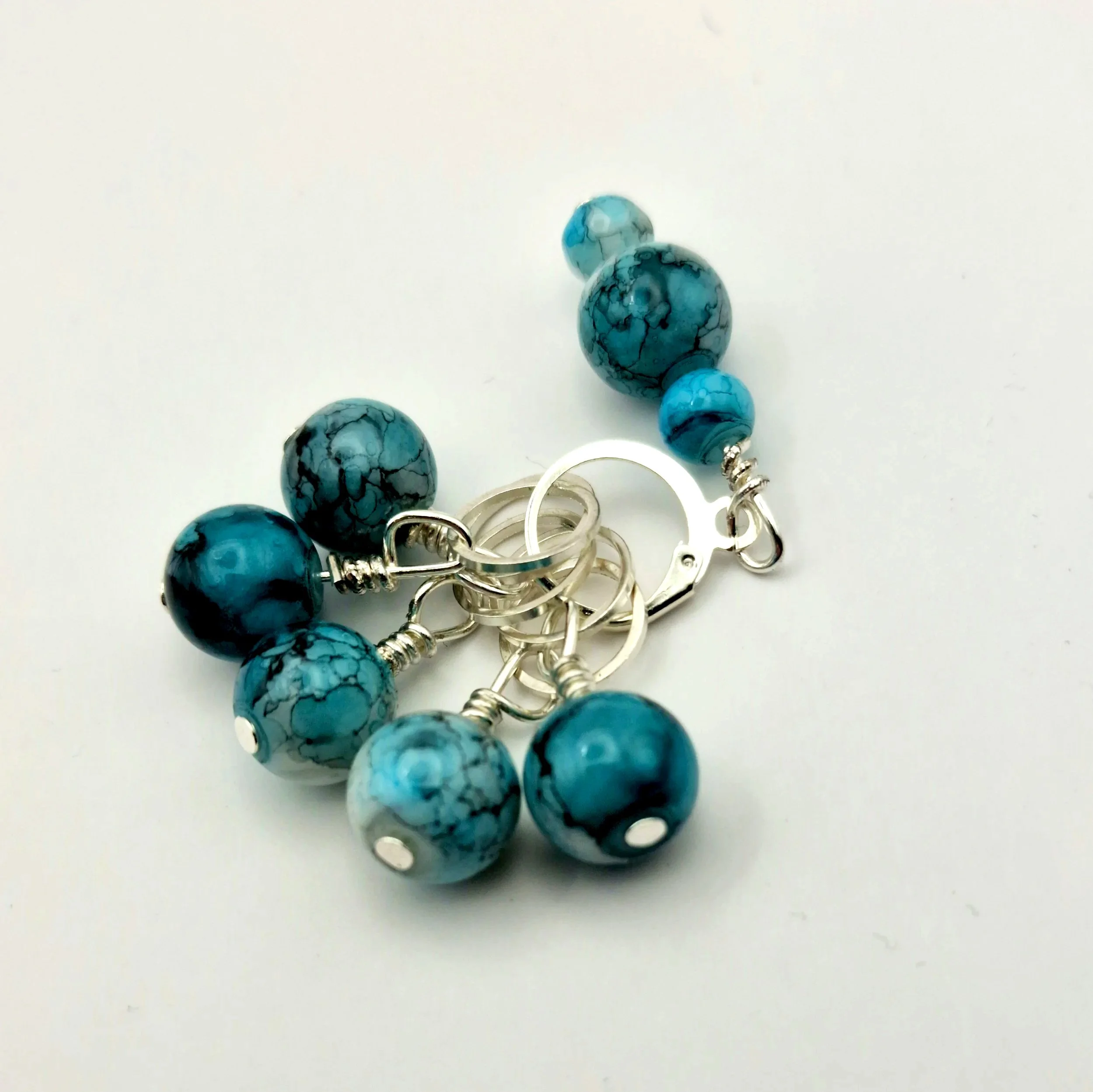 Set aus fünf Maschenmarkierern und einem Progresskeeper aus Teal und türkisfarbenen handbemalten Keramikperlen kombiniert mit versilberten Ringen auf weißem Untergrund