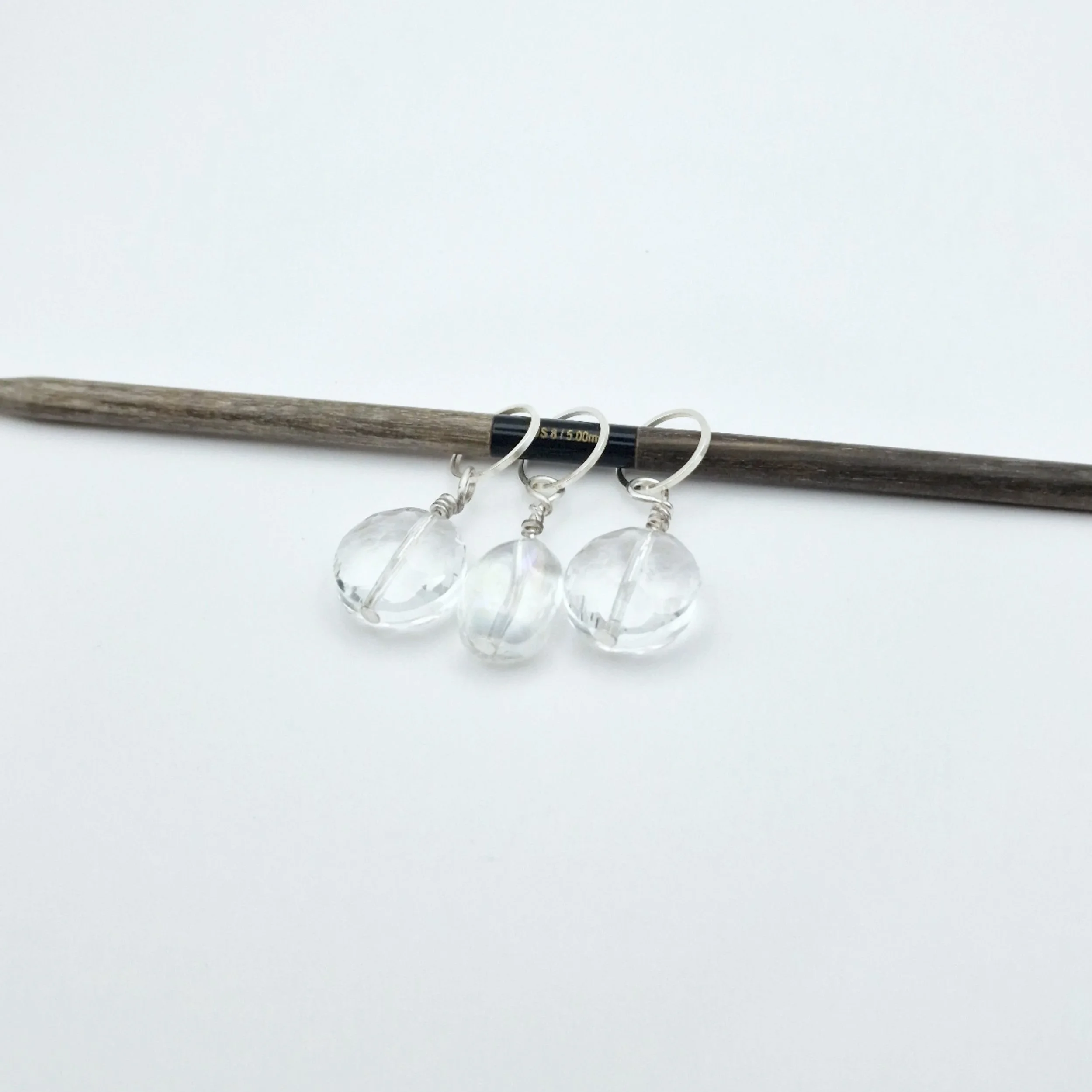 Drei Maschenmarkierer aus klarem Glas mit silberfarbenem Metall auf weißem Untergrund und einer 5 mm Stricknadel aus Holz.