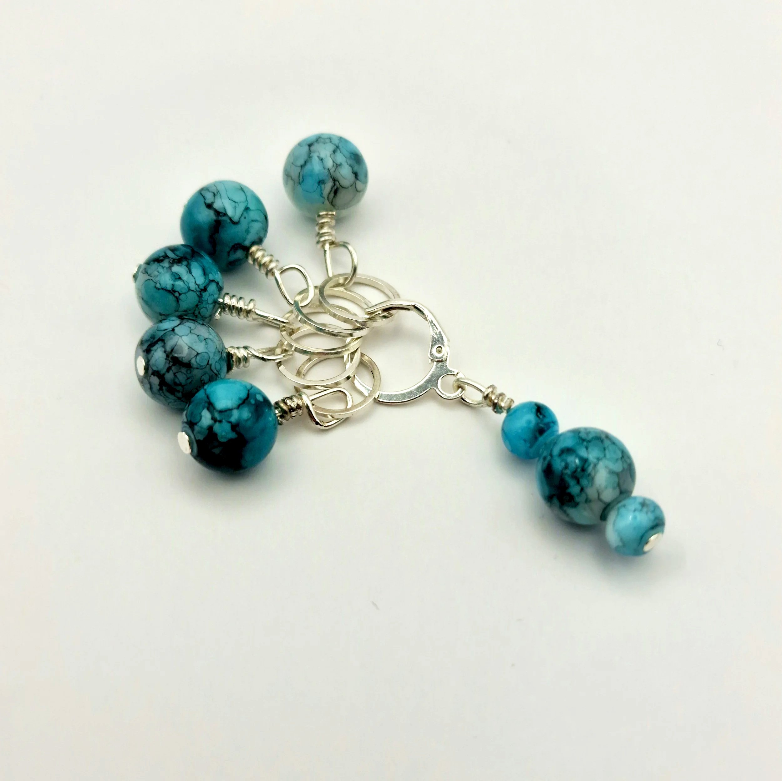 Set aus fünf Maschenmarkierern und einem Progresskeeper aus Teal und türkisfarbenen handbemalten Keramikperlen kombiniert mit versilberten Ringen auf weißem Untergrund