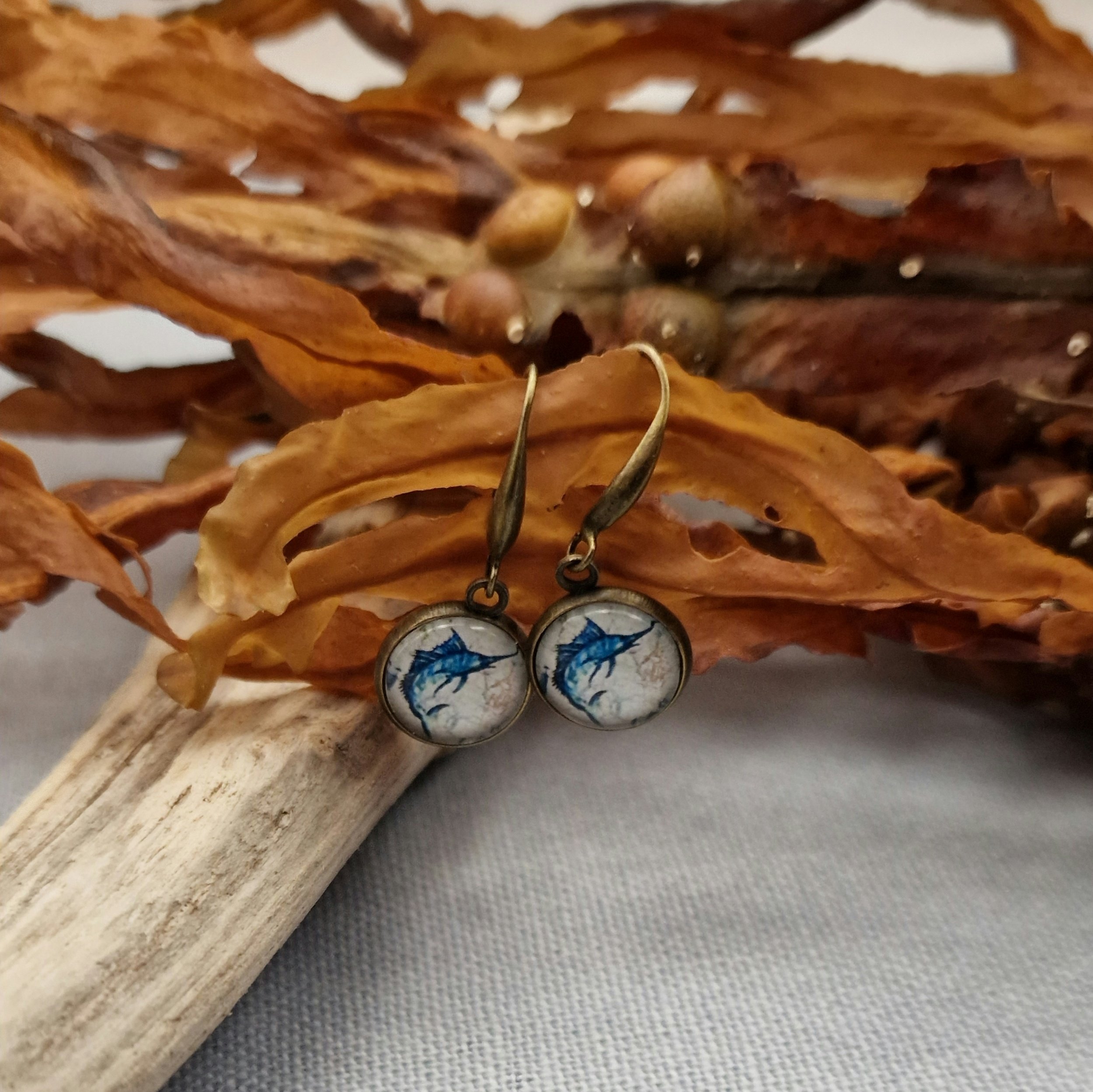 ein paar hängende Ohrringe in Bronze mit Cabochon mit Bild von Schwertfisch hängen an getrocknetem Seetang und Treibholz auf hellblauem Untergrund