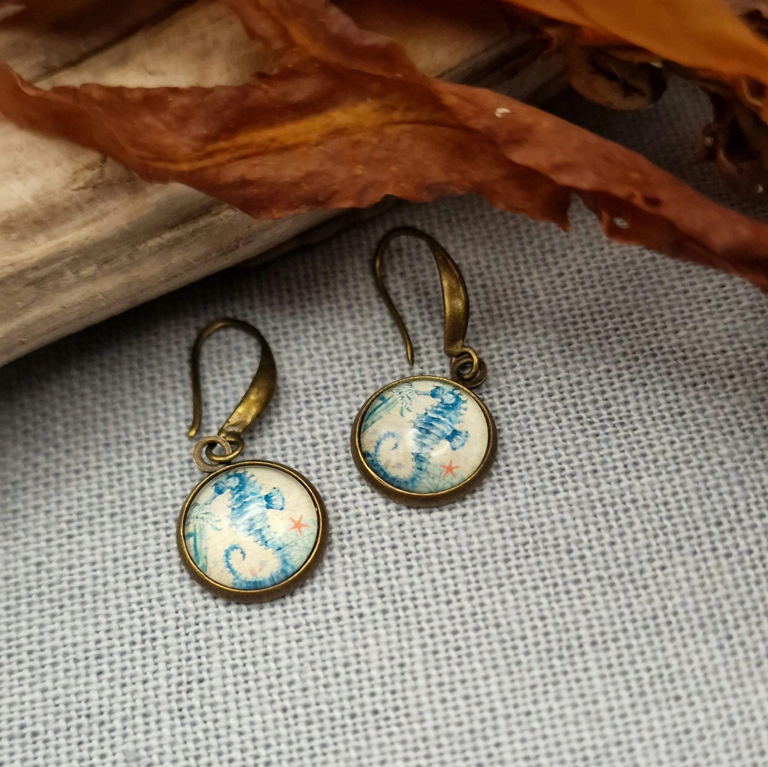 ein paar hängende Ohrringe in Bronze mit Cabochon mit Bild von Seepferdchen liegen auf auf hellblauem Untergrund