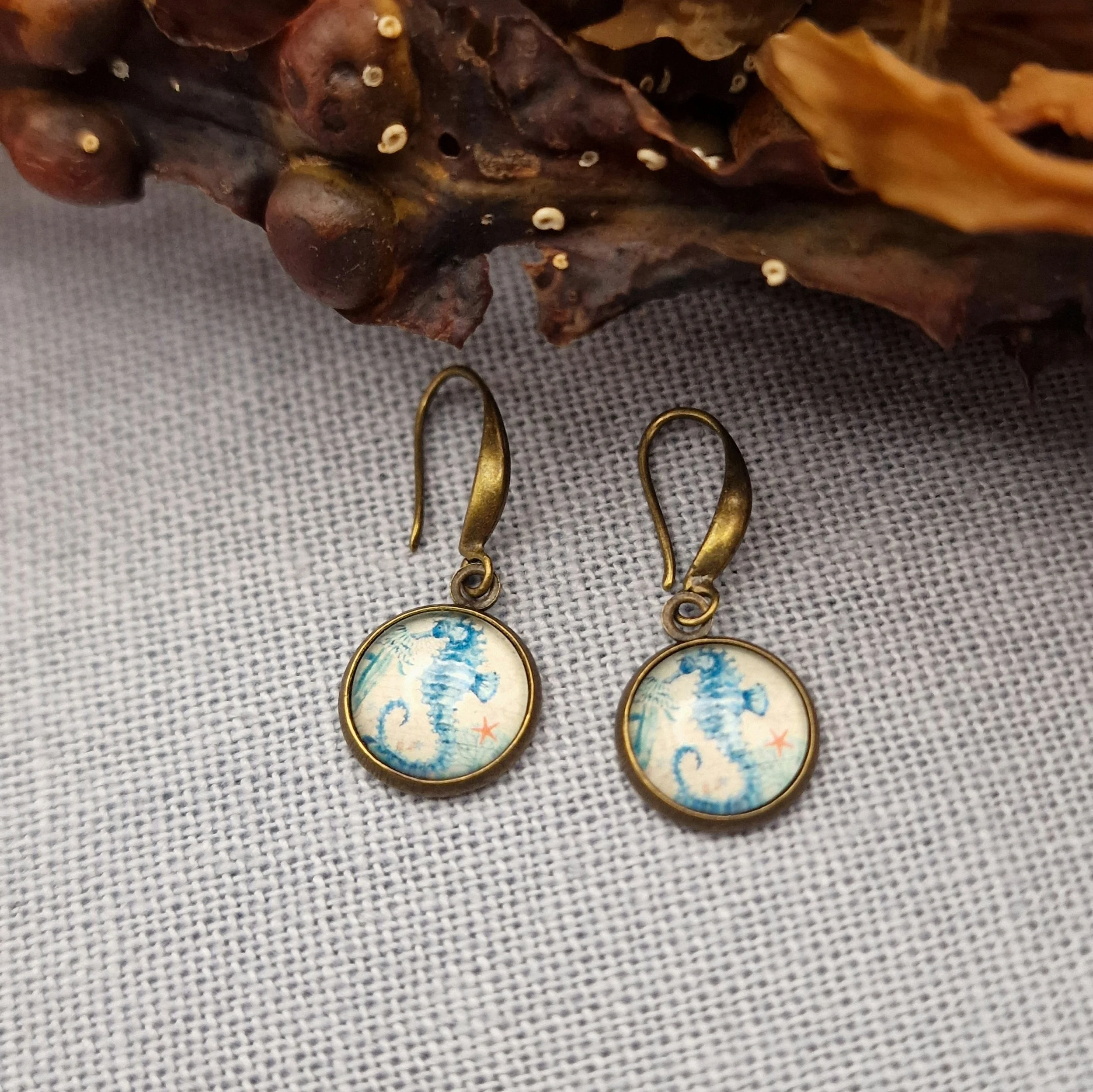 ein paar hängende Ohrringe in Bronze mit Cabochon mit Bild von Seepferdchen liegen auf auf hellblauem Untergrund