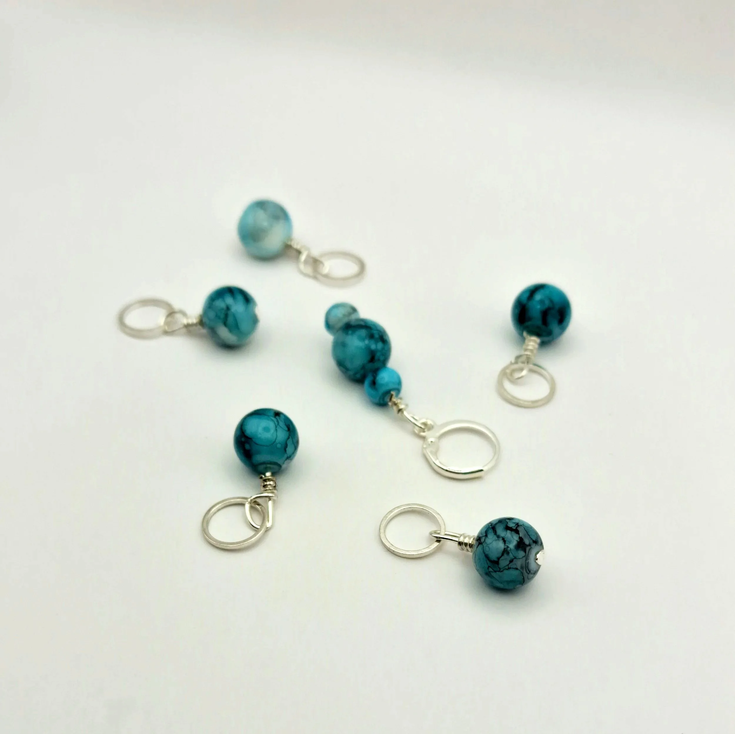 Set aus fünf Maschenmarkierern und einem Progresskeeper aus Teal und türkisfarbenen handbemalten Keramikperlen kombiniert mit versilberten Ringen auf weißem Untergrund