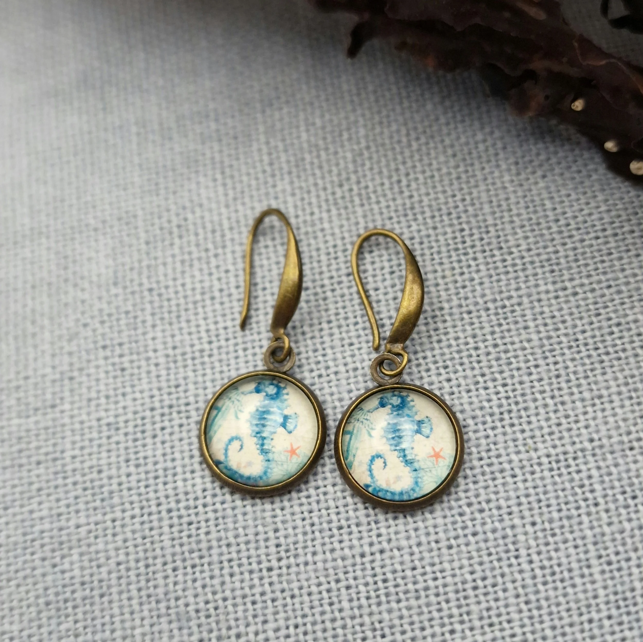 ein paar hängende Ohrringe in Bronze mit Cabochon mit Bild von Seepferdchen liegen auf auf hellblauem Untergrund