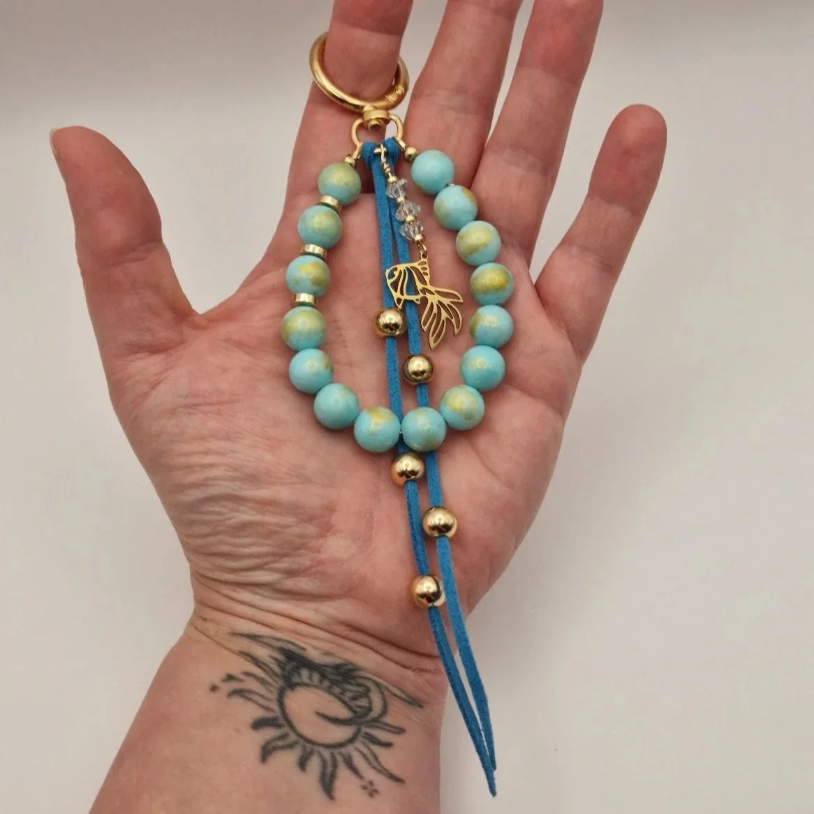 handgefertigter Taschenanhänger Smáfiskur mit türkisfarbenen Natursteinperlen, vergoldeten Hämatit - Discs, blauem Wildlederband und Fisch Charm aus vergoldetem Edelstahl auf einer Hand liegend