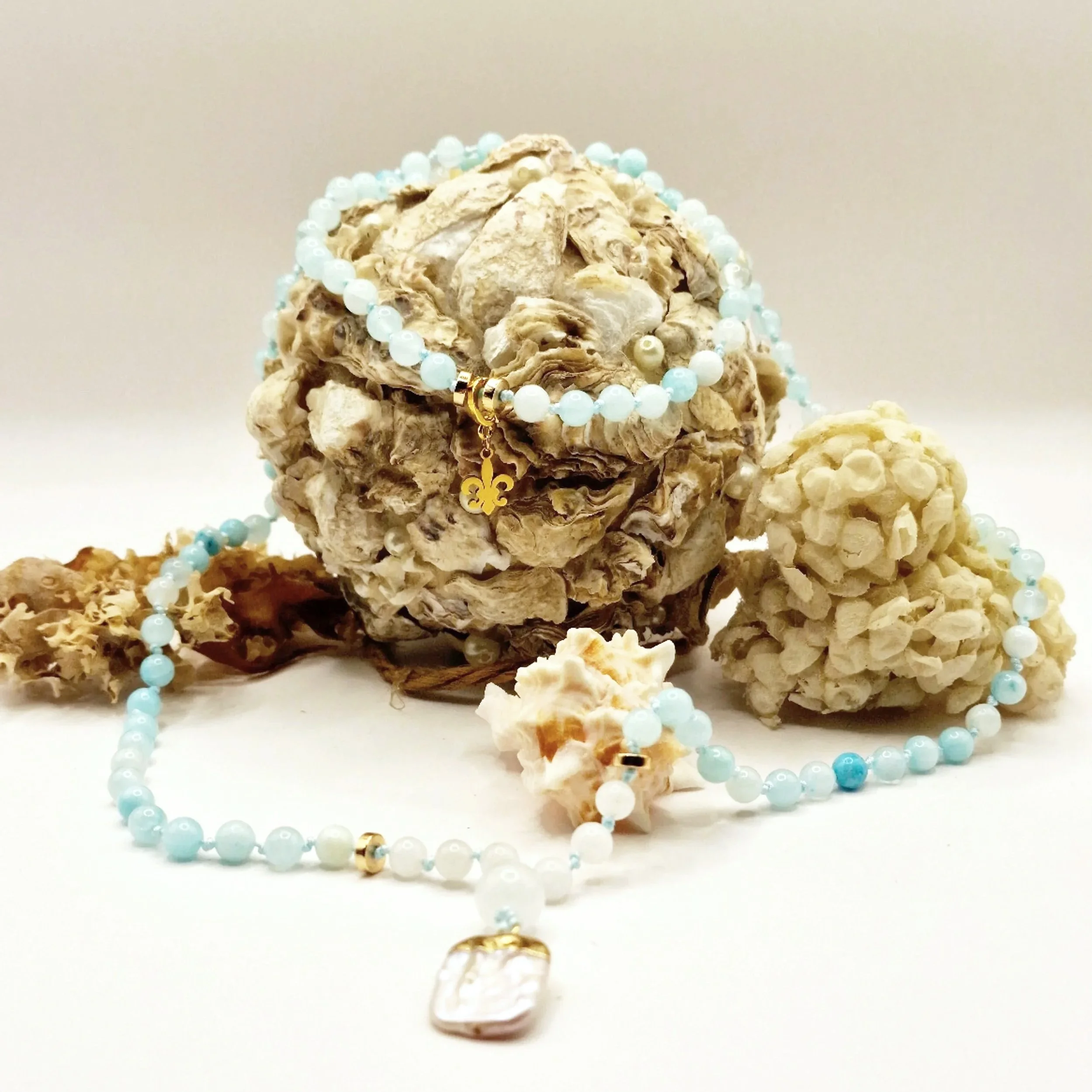 Kette aus hellblauen Natursteinperlen mit Süßwasserperle als Anhänger und goldfarbenen Akzenten dekoriert mit Muscheln auf hellem Untergrund