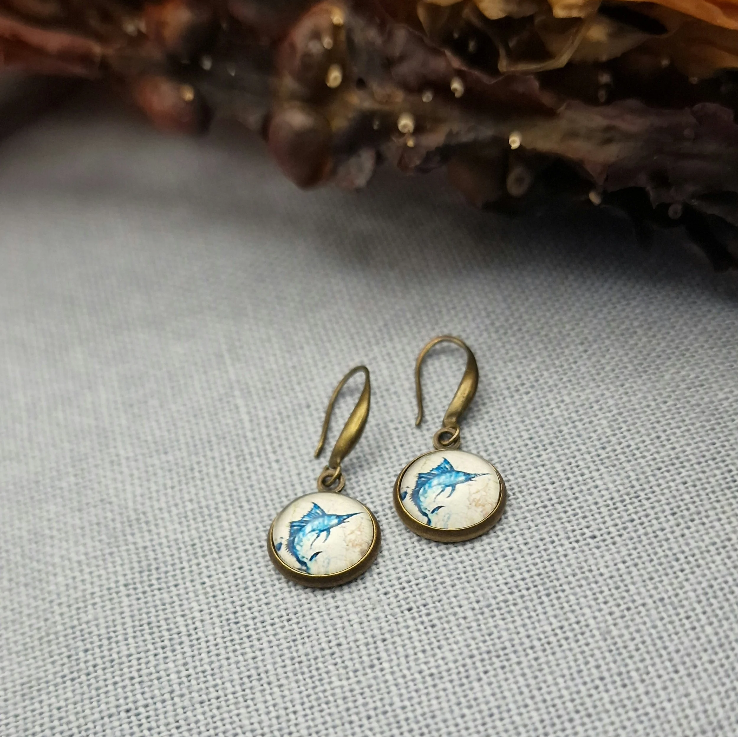 ein paar hängende Ohrringe in Bronze mit Cabochon mit Bild von Schwertfisch hängen an getrocknetem Seetang und Treibholz auf hellblauem Untergrund