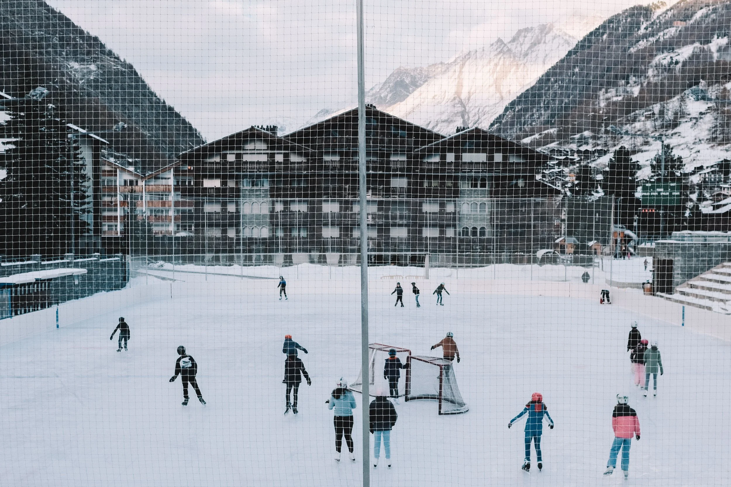 People playing hockey on an outdoor ice rink surrounded by snow-covered buildings and mountains.