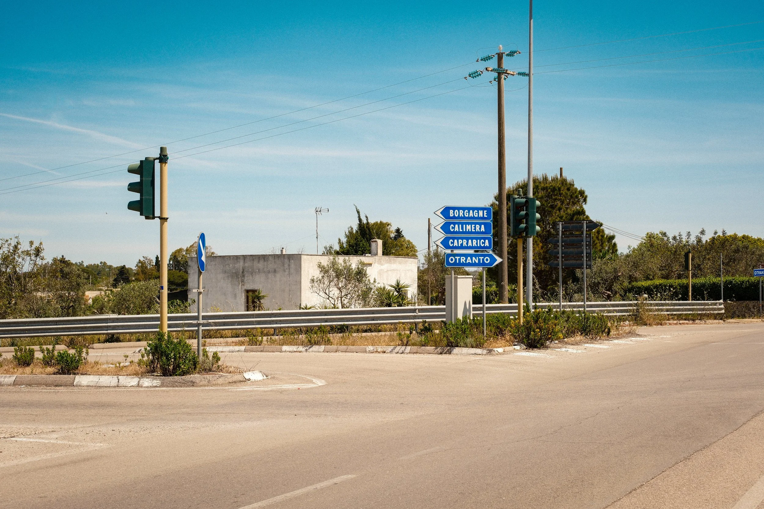 A road intersection with blue directional signs pointing to Borgagne, Calimera, Caprarica, and Otranto. Traffic lights and utility poles are also visible under a clear blue sky.