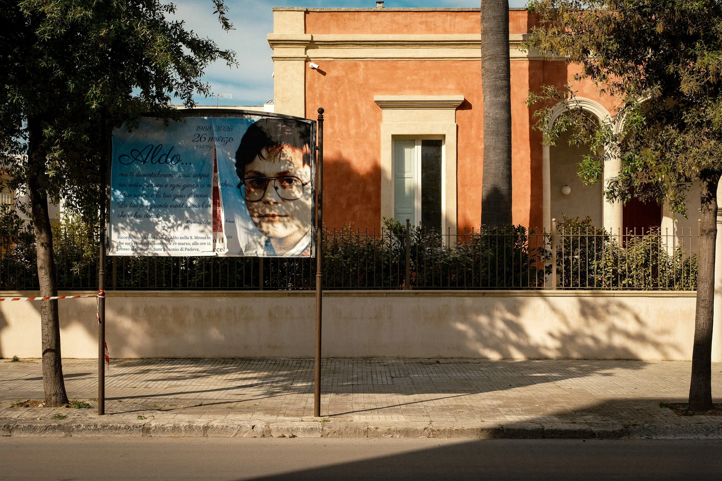 A billboard with a portrait of a young man wearing glasses and text in Italian, located in front of a salmon-colored building with trees and a fence in the foreground.