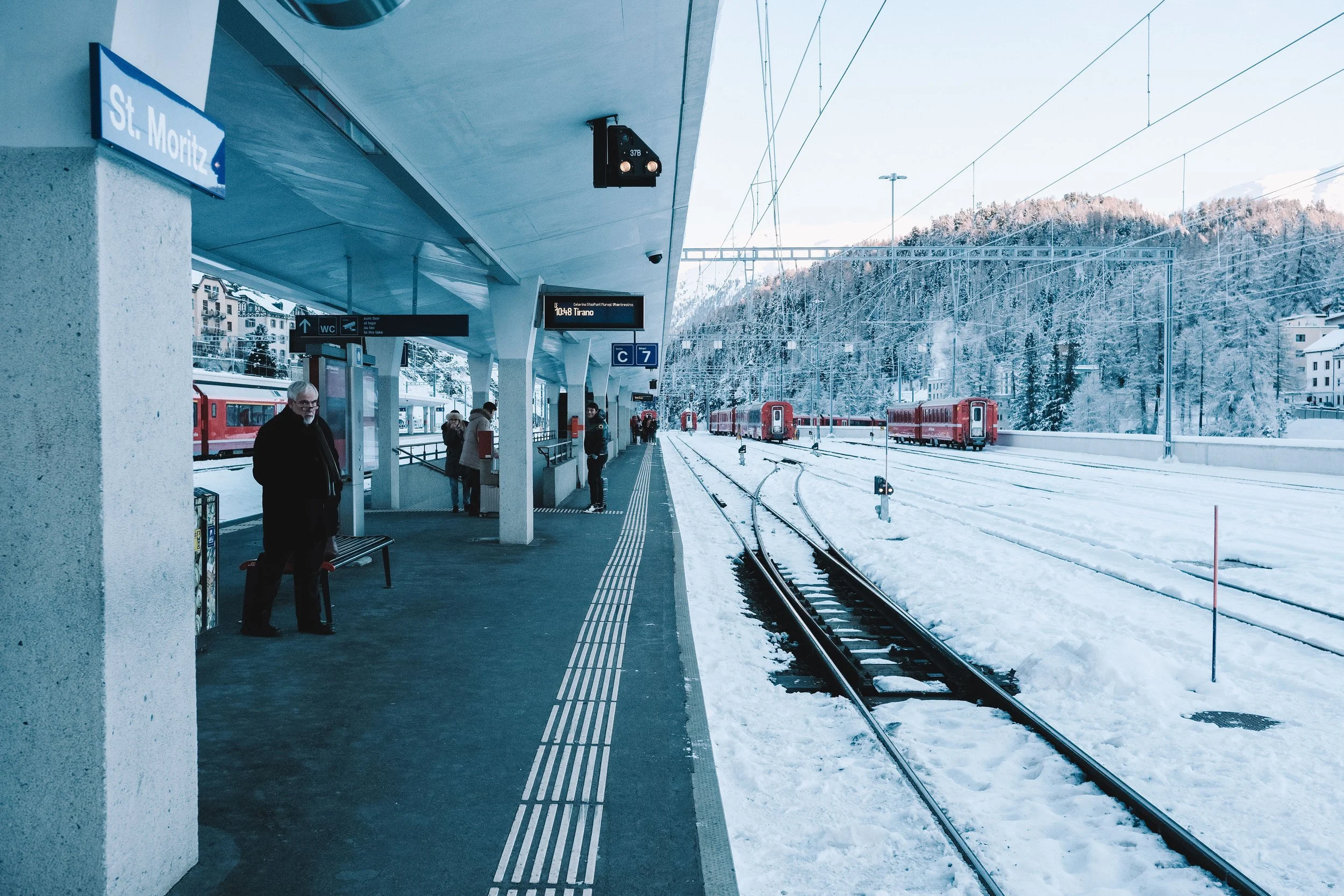 St. Moritz train station covered in snow with red trains and people on platform.